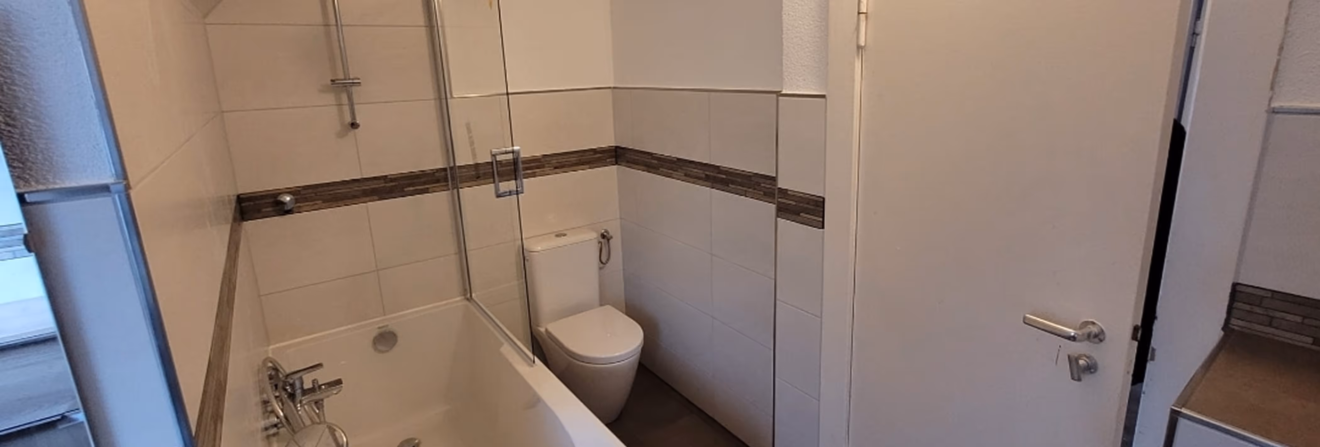 Kleines Badezimmer mit Badewanne, Duschwand aus Glas, Toilette und weißen Wandfliesen mit braunem horizontalem Muster.