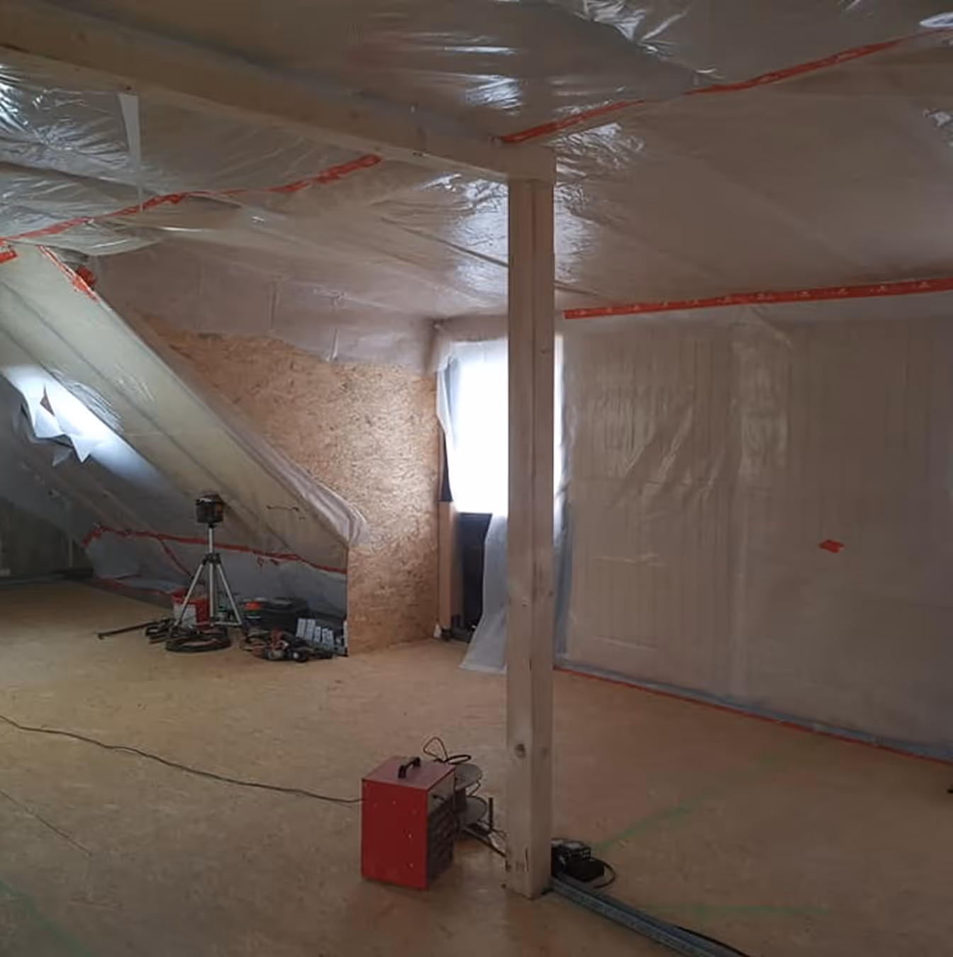 Ausgebauter Dachboden mit Holzstütze, Boden aus OSB-Platten und Plastikfolie an Wänden und Decke, Bauwerkzeuge verteilt.