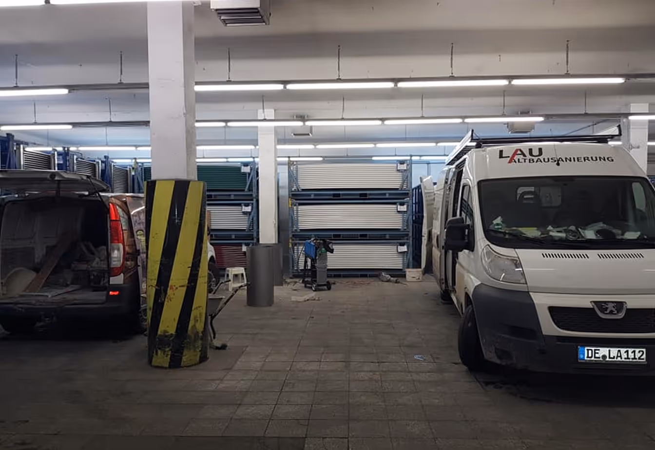 Parkhaus mit zwei geparkten Transportern auf beiden Seiten eines mittigen Pfeilers mit gelb-schwarzen Warnstreifen, einem Lagerregal mit weißen Platten und mehreren Werkzeugen auf dem Boden.