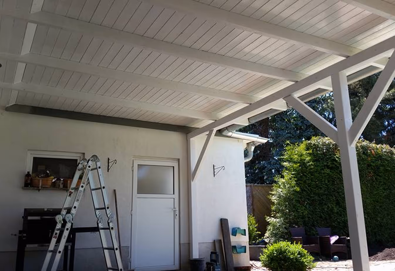 Überdachte Terrasse mit weißem Holzdeckendach, weißer Tür, Leiter und Gartenmöbeln im Hintergrund.