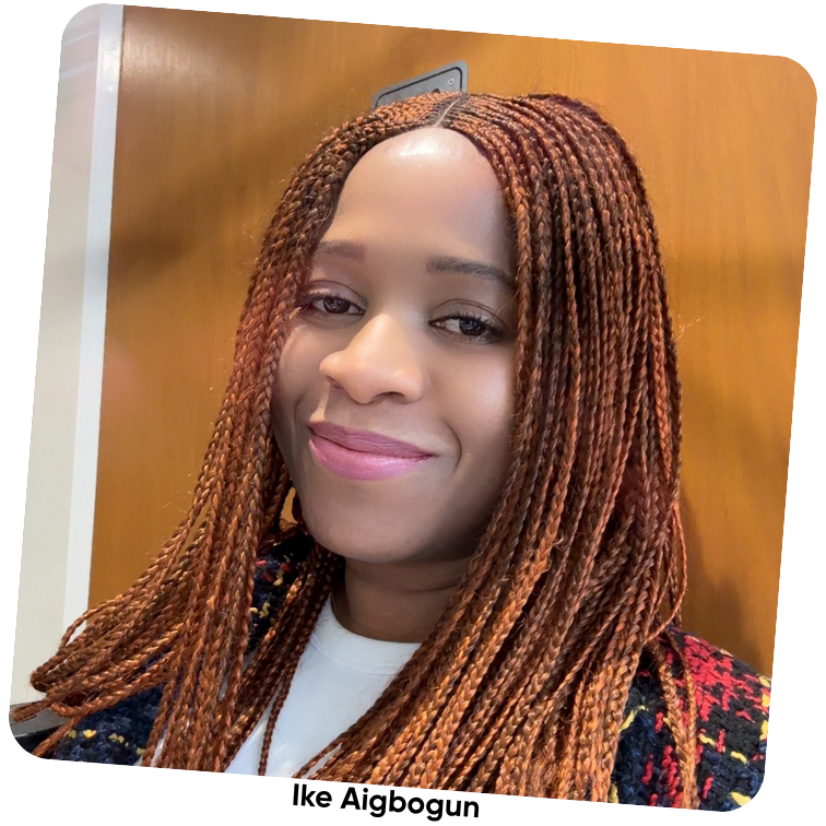 Ike - Founder of VIGIL. Picture of Smiling woman with long reddish-brown braided hair wearing a patterned jacket and white top, standing indoors with a wooden door in the background.
