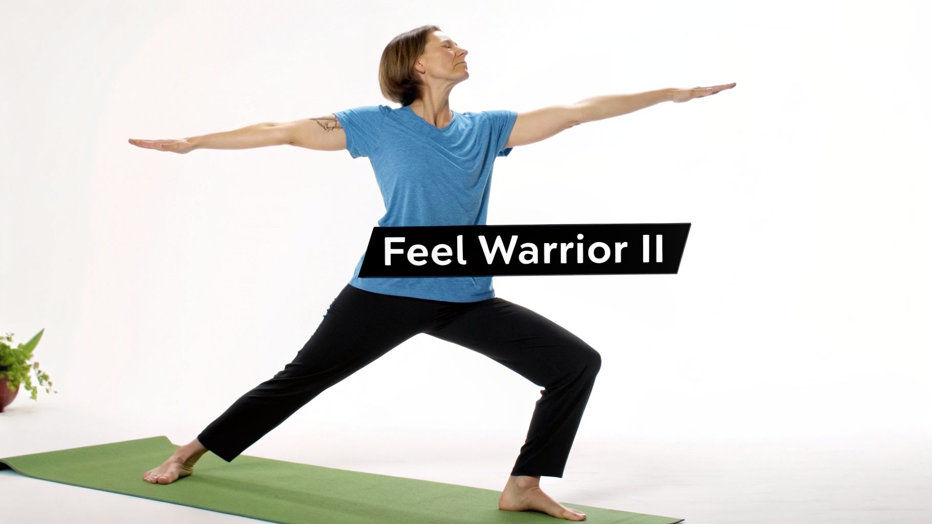A woman in a blue shirt and black pants practices Warrior II yoga pose on a green mat.