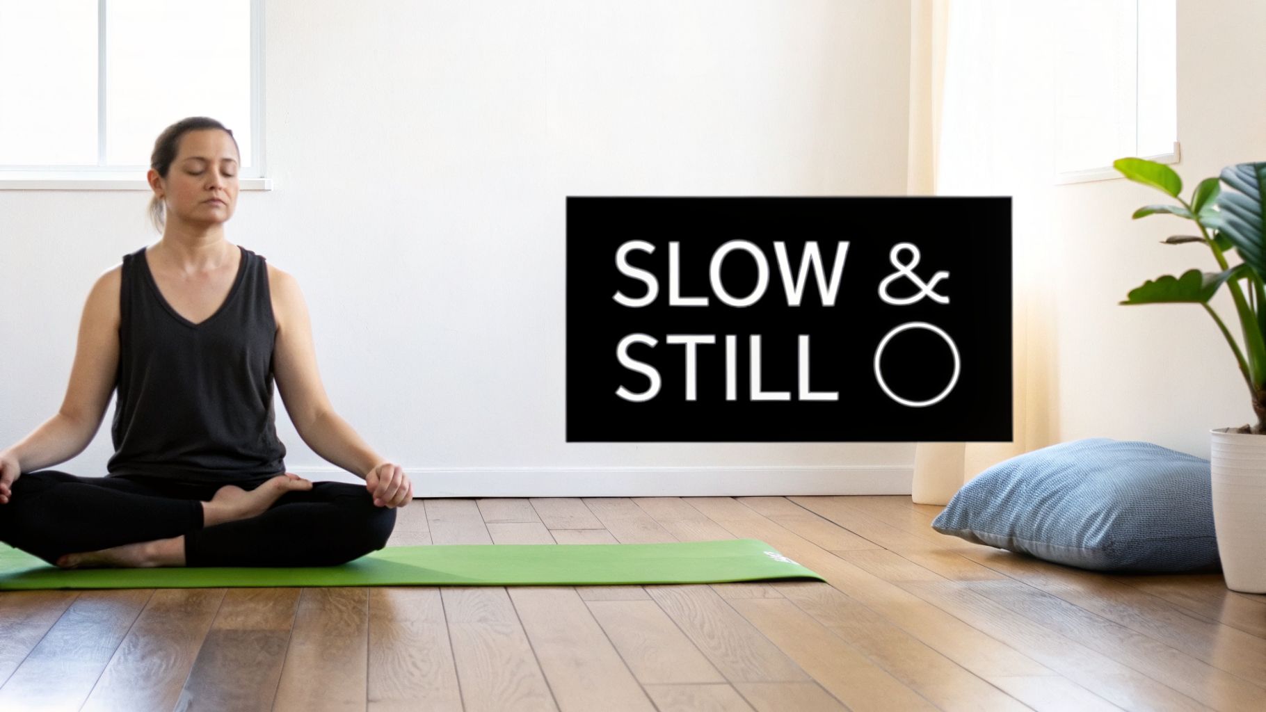 A serene woman meditating in a cross-legged yoga pose on a green mat in a bright room.
