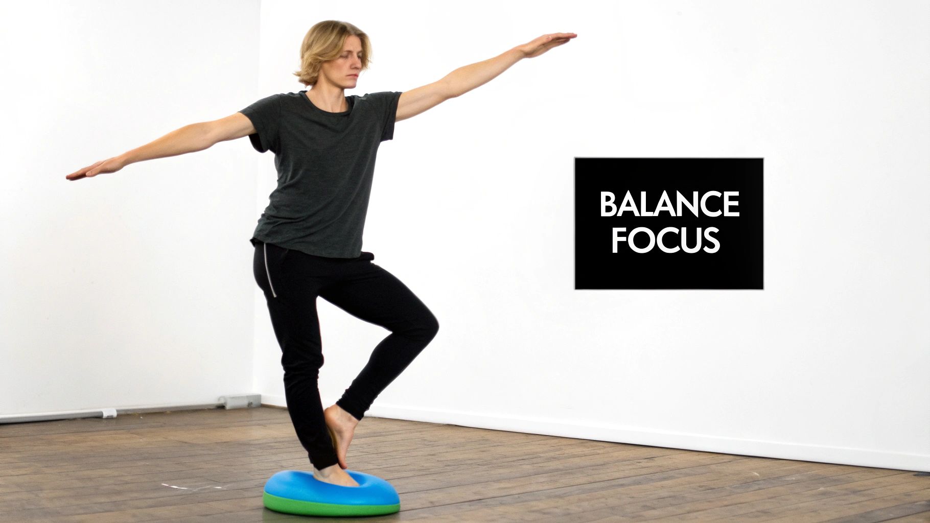 A man balances on one leg on a blue and green stability cushion, arms extended, on a wooden floor.