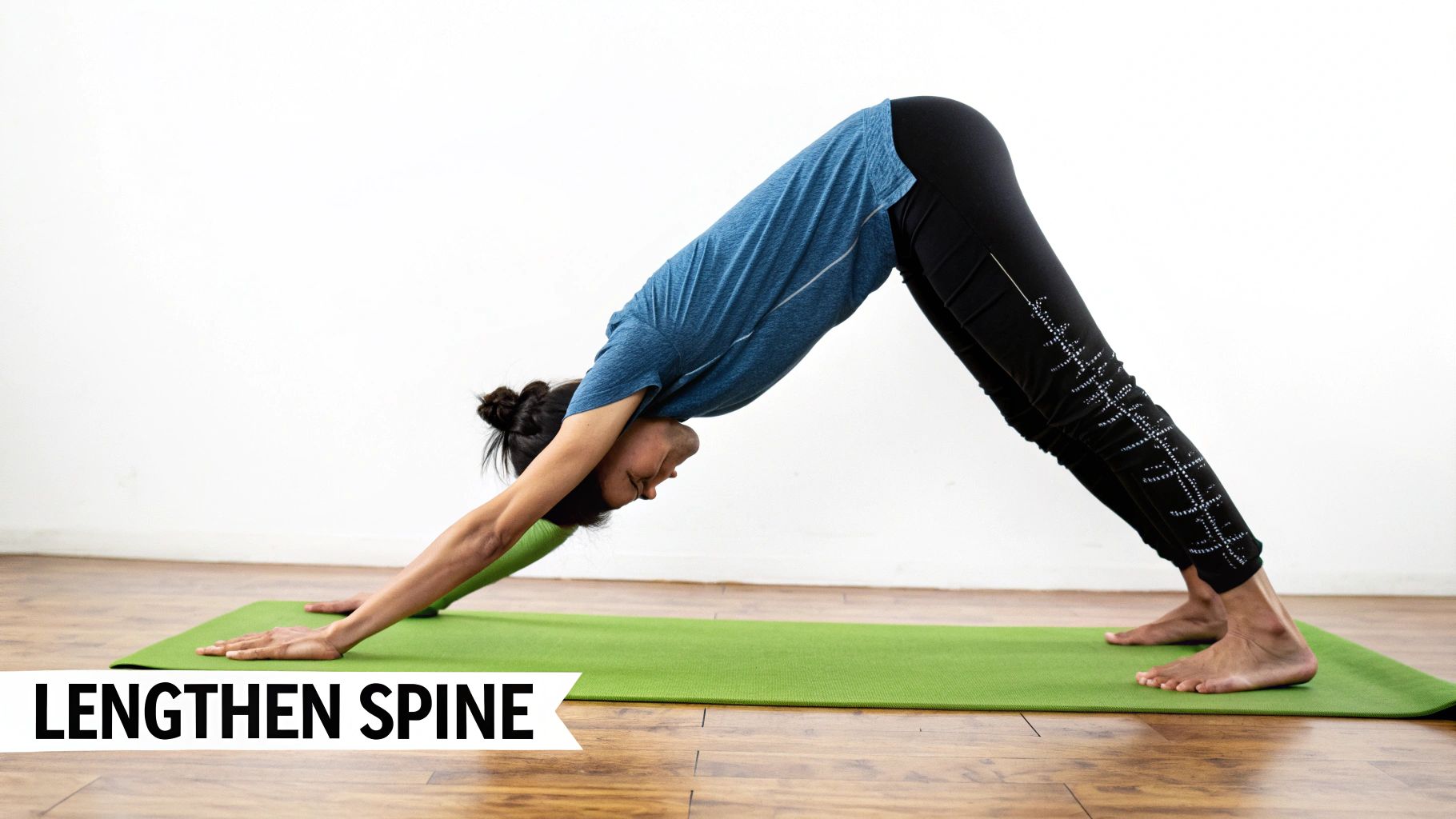 A woman in a blue shirt and black leggings performs downward-facing dog yoga on a green mat to lengthen her spine.
