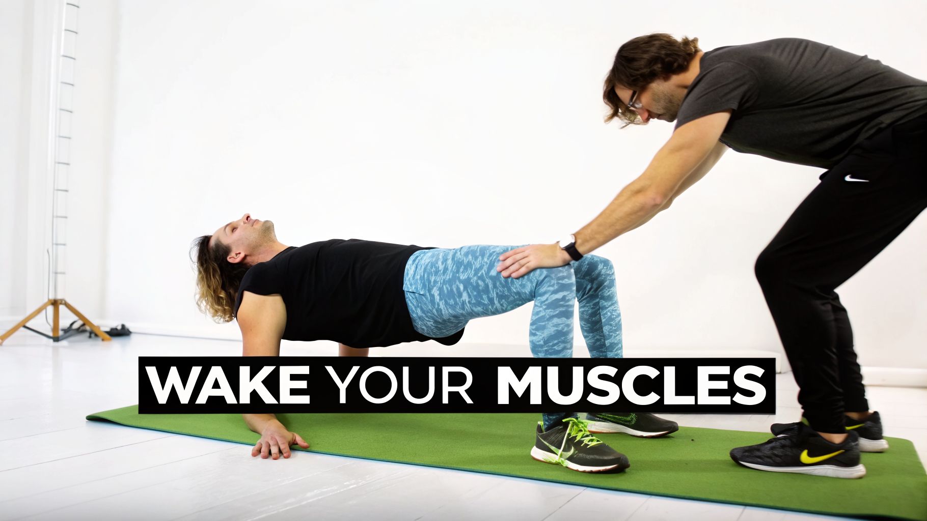 Two men exercise on a mat, one performing a bridge pose with assistance from a trainer, promoting muscle activation.