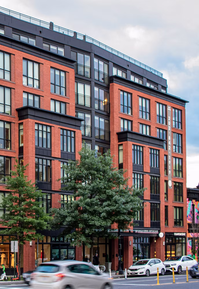 Modern red brick commercial building with large windows, street trees, and cars driving by.