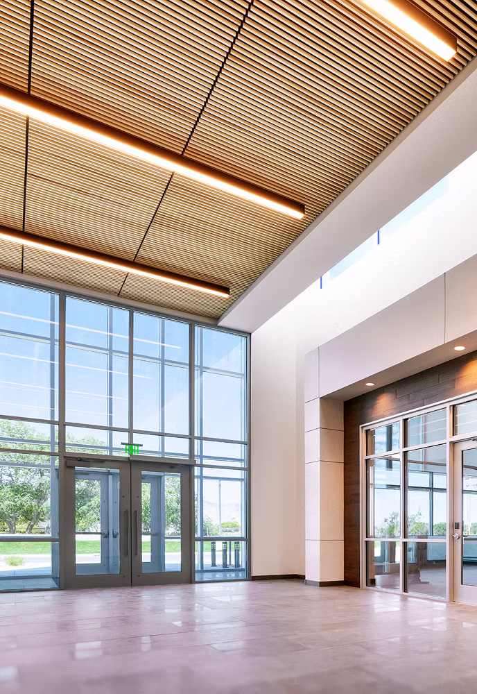 Spacious modern building interior with large glass windows, wooden slatted ceiling panels, and tile flooring.