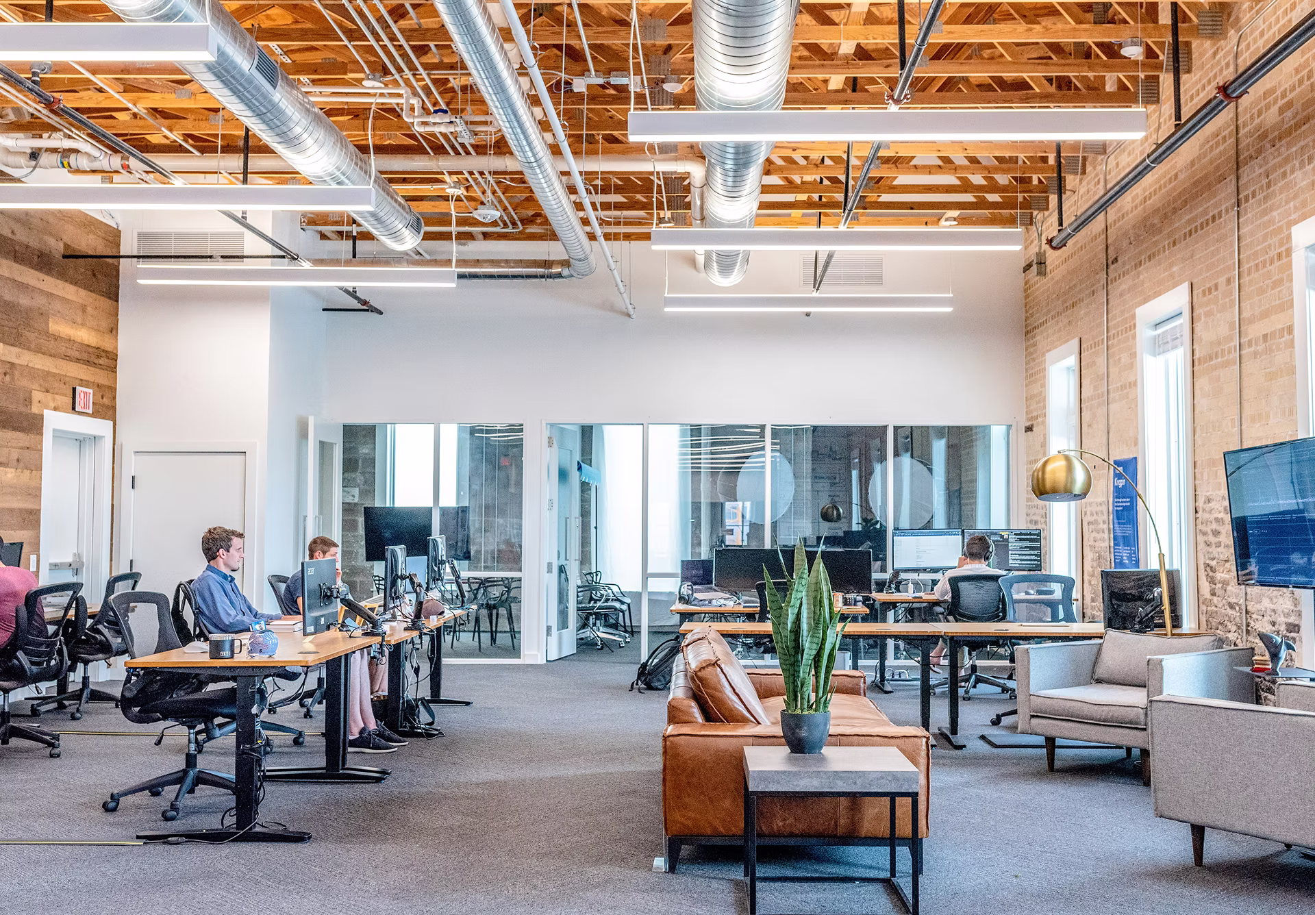 Modern office space with people working at desks, exposed wooden ceiling beams, and a seating area with a leather couch and gray chairs.