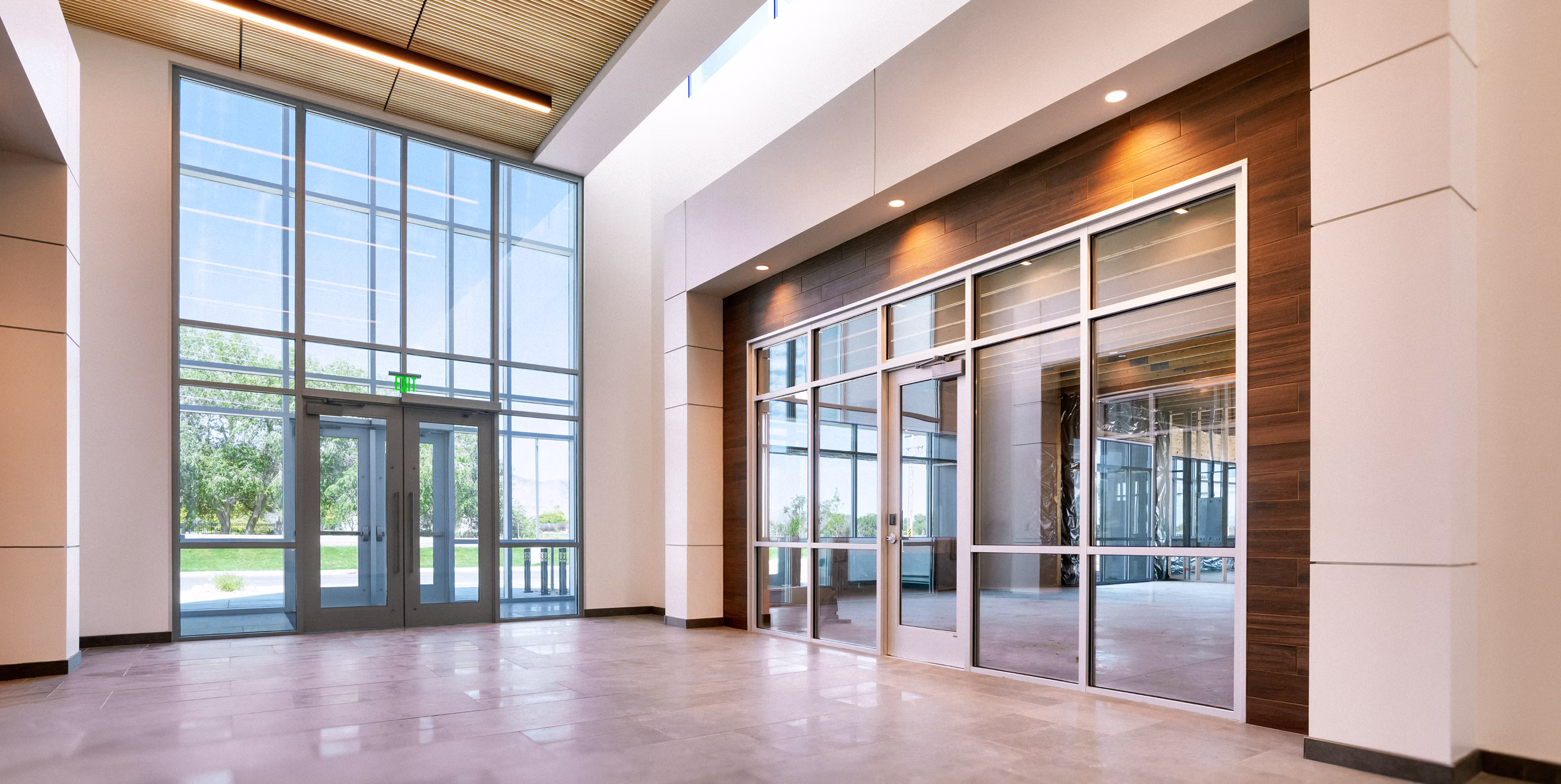 Bright modern lobby with large glass entrance doors and a wall of floor-to-ceiling windows next to a wooden-paneled glass office door.