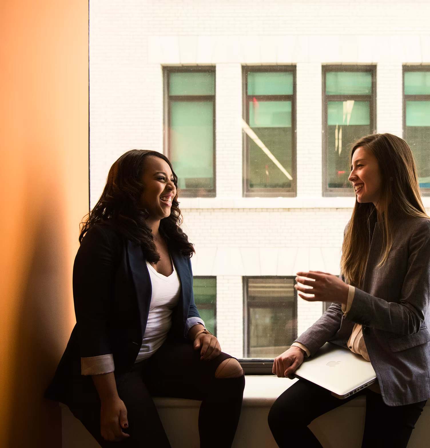 Two professional women laughing and talking while sitting on a window sill in a bright, modern office with a city building visible through the glass.