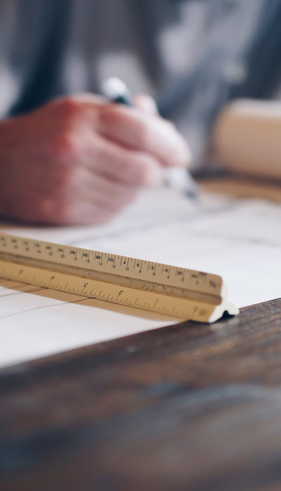 Close-up of a wooden triangular ruler on a table with a person sketching on paper in the background.