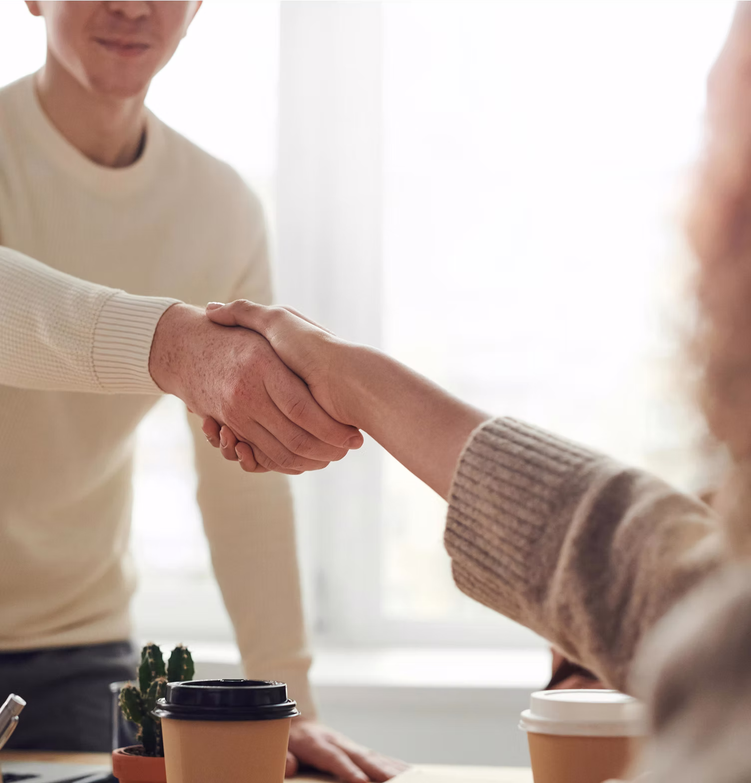 Two people shaking hands over a table with coffee cups and a small cactus plant.
