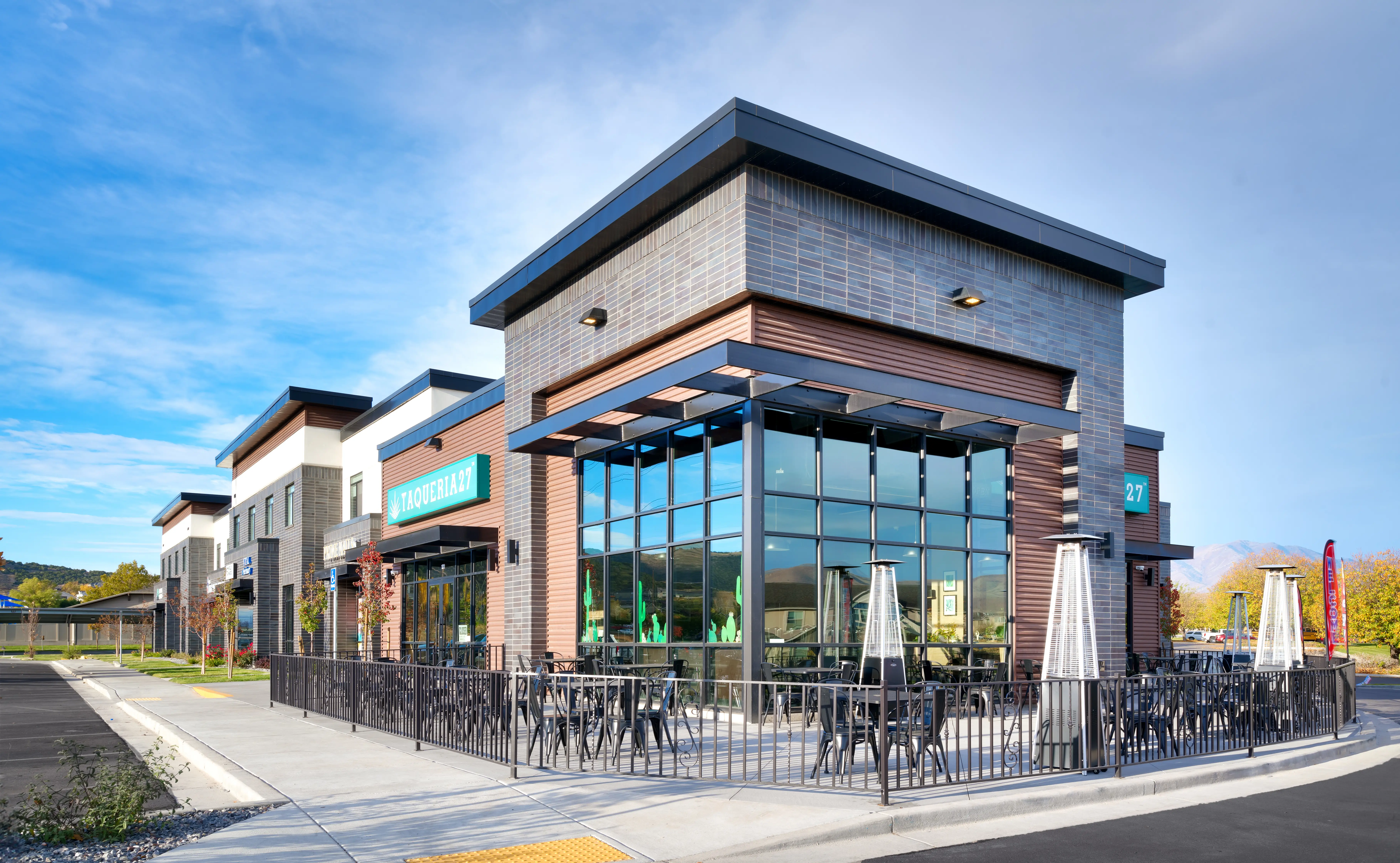 Modern corner restaurant Taqueria 27 with large glass windows, outdoor seating area, and patio heaters under a blue sky.