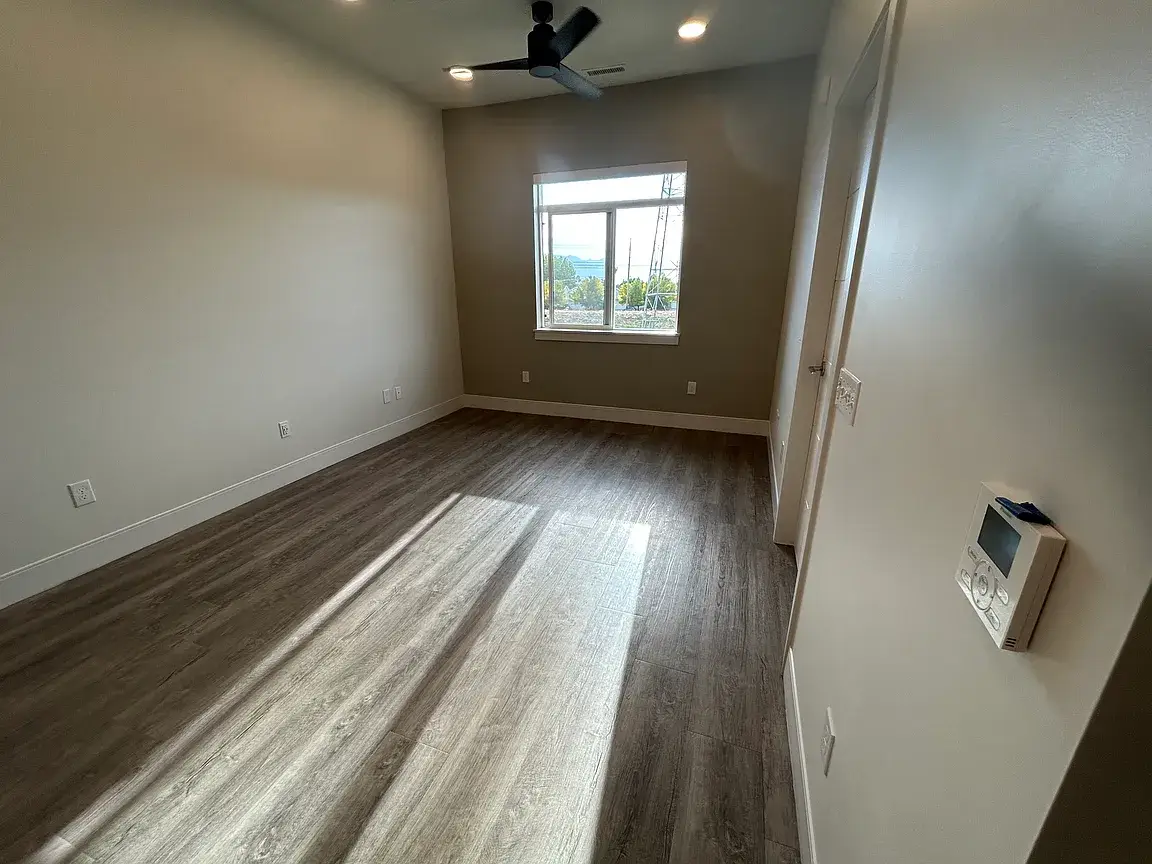 Interior view of a modern, empty bedroom featuring light grey walls, dark wood-style plank flooring, a sleek black ceiling fan, and bright natural light streaming from a large window.