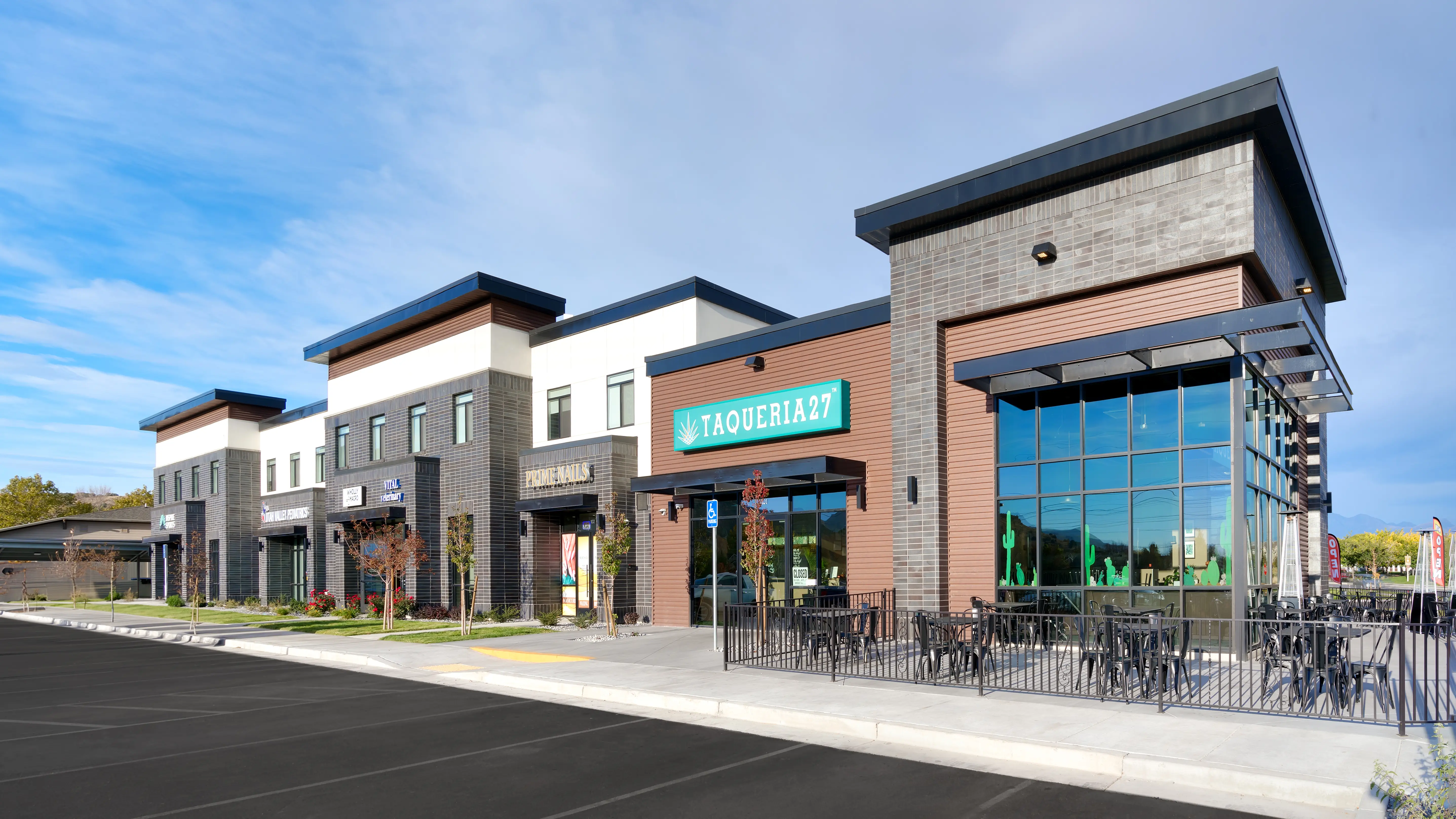 Modern commercial building with multiple storefronts including Taqueria 27 and Prime Nails, with outdoor seating area and clear blue sky.