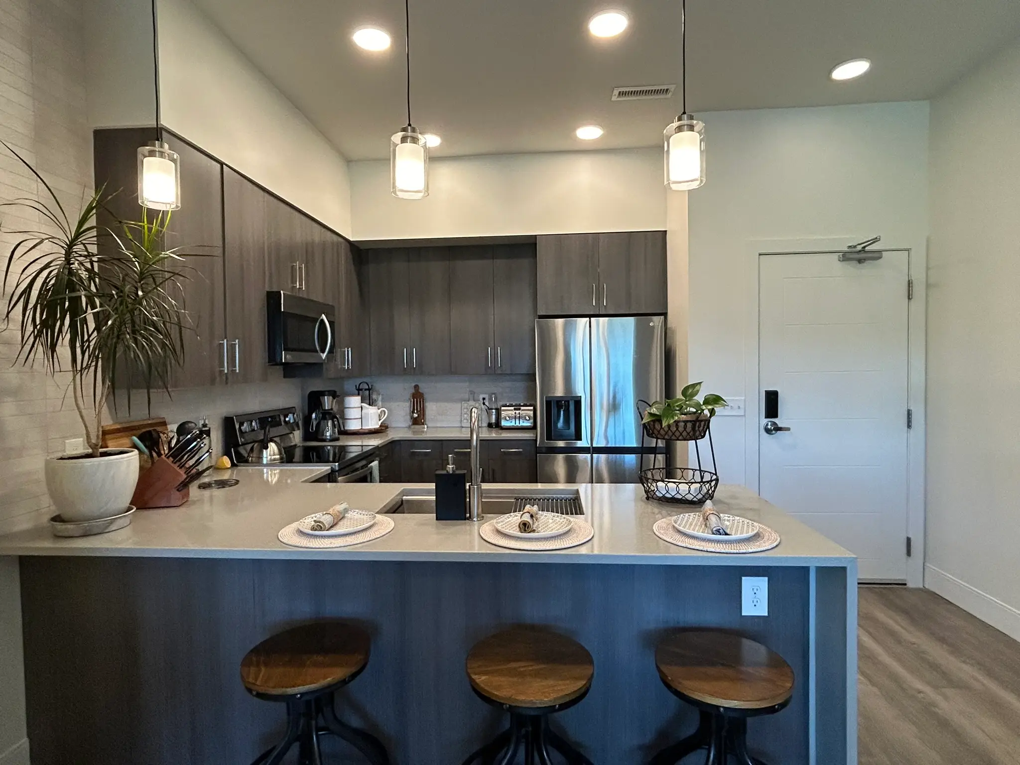 Modern kitchen with gray cabinets, stainless steel appliances, three wooden stools, and a countertop set with plates and napkins.