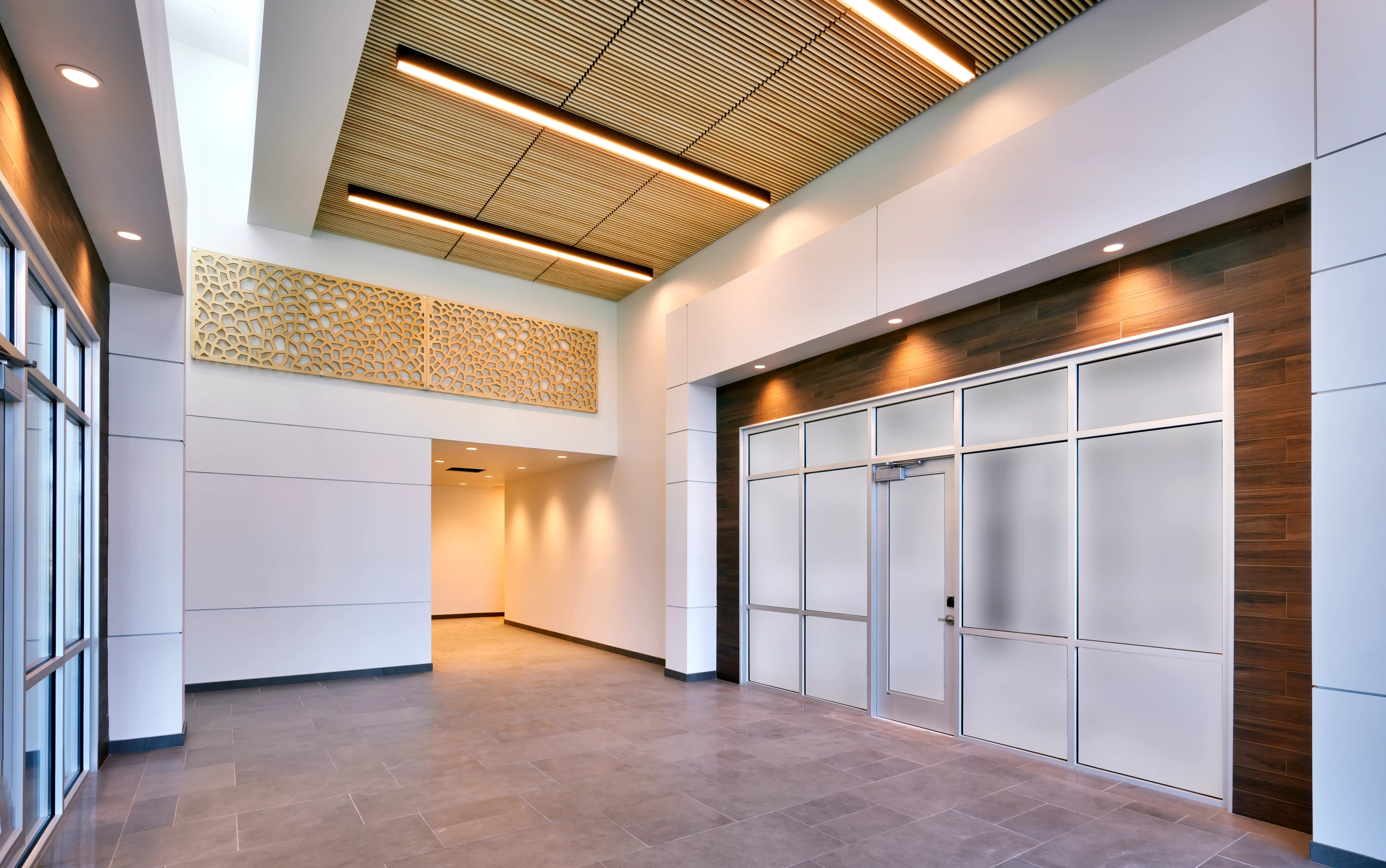 Modern interior hallway with wood-paneled wall, frosted glass doors, tiled floor, and decorative wooden ceiling panels with lighting.