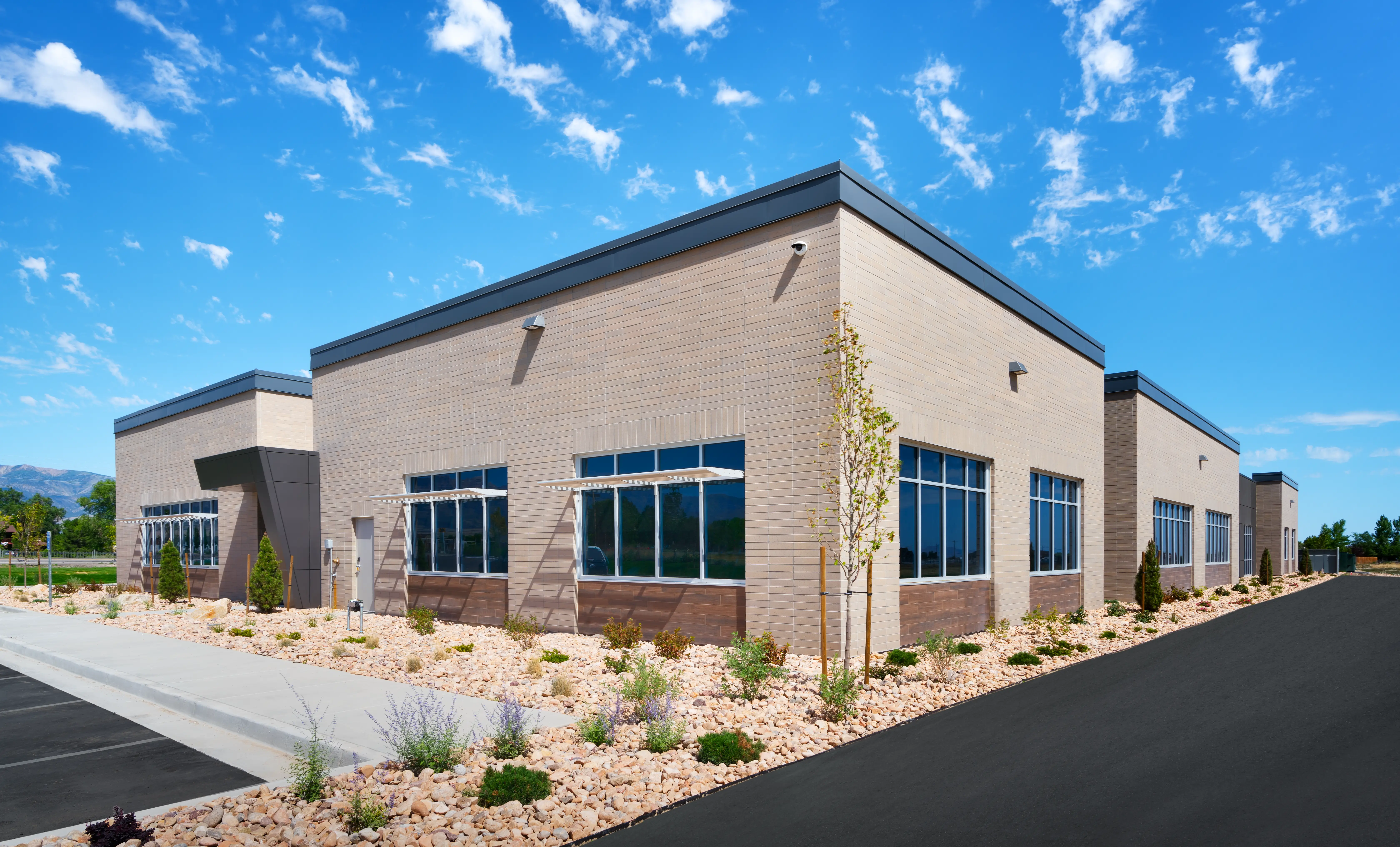 Modern beige brick commercial building under blue sky with landscaped rock garden and shrubs along sidewalk.