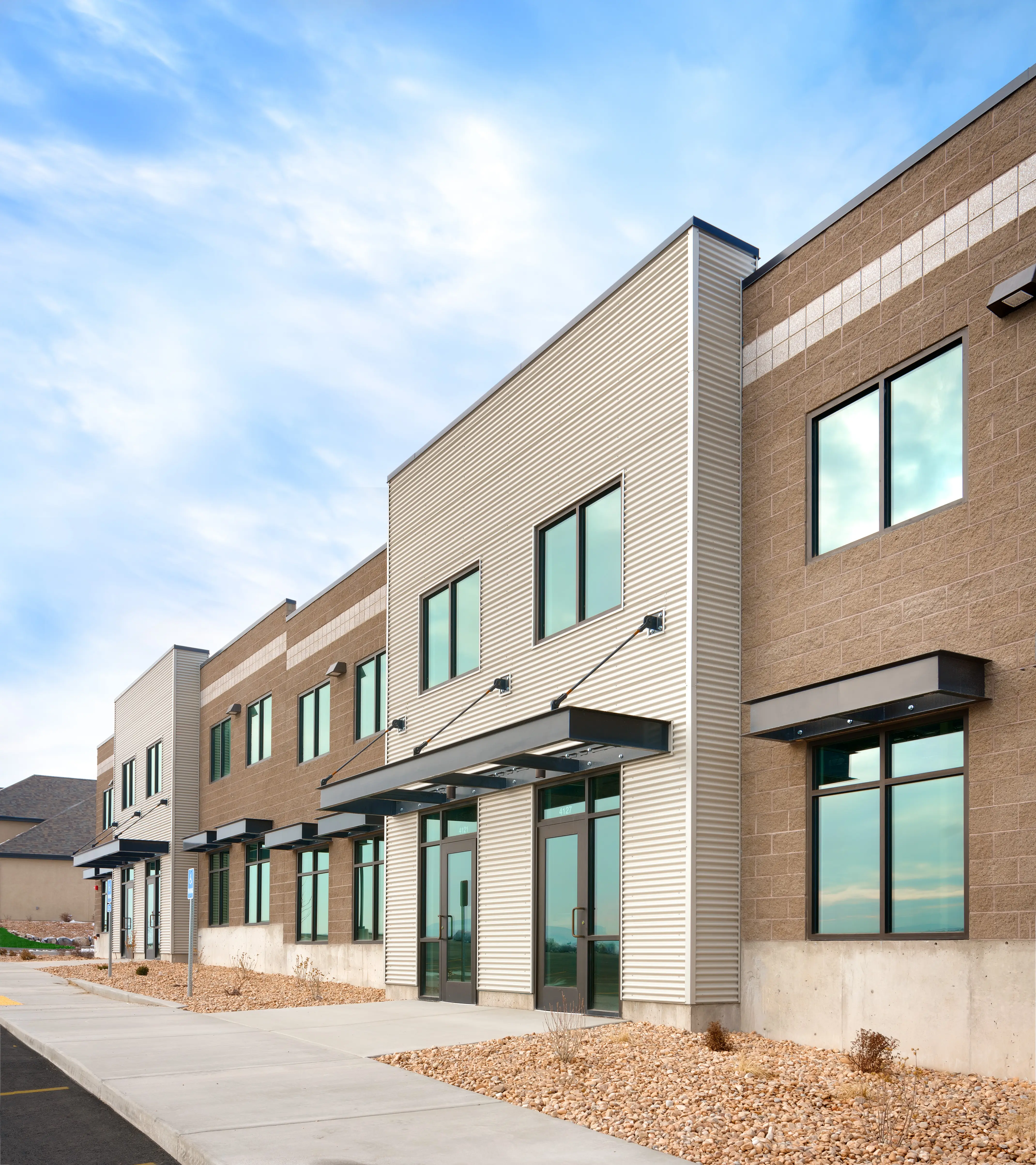 Exterior view of a modern industrial-style commercial building featuring tan stone masonry and vertical white corrugated metal siding. Large glass storefront doors sit under sleek black metal awnings against a bright blue sky.