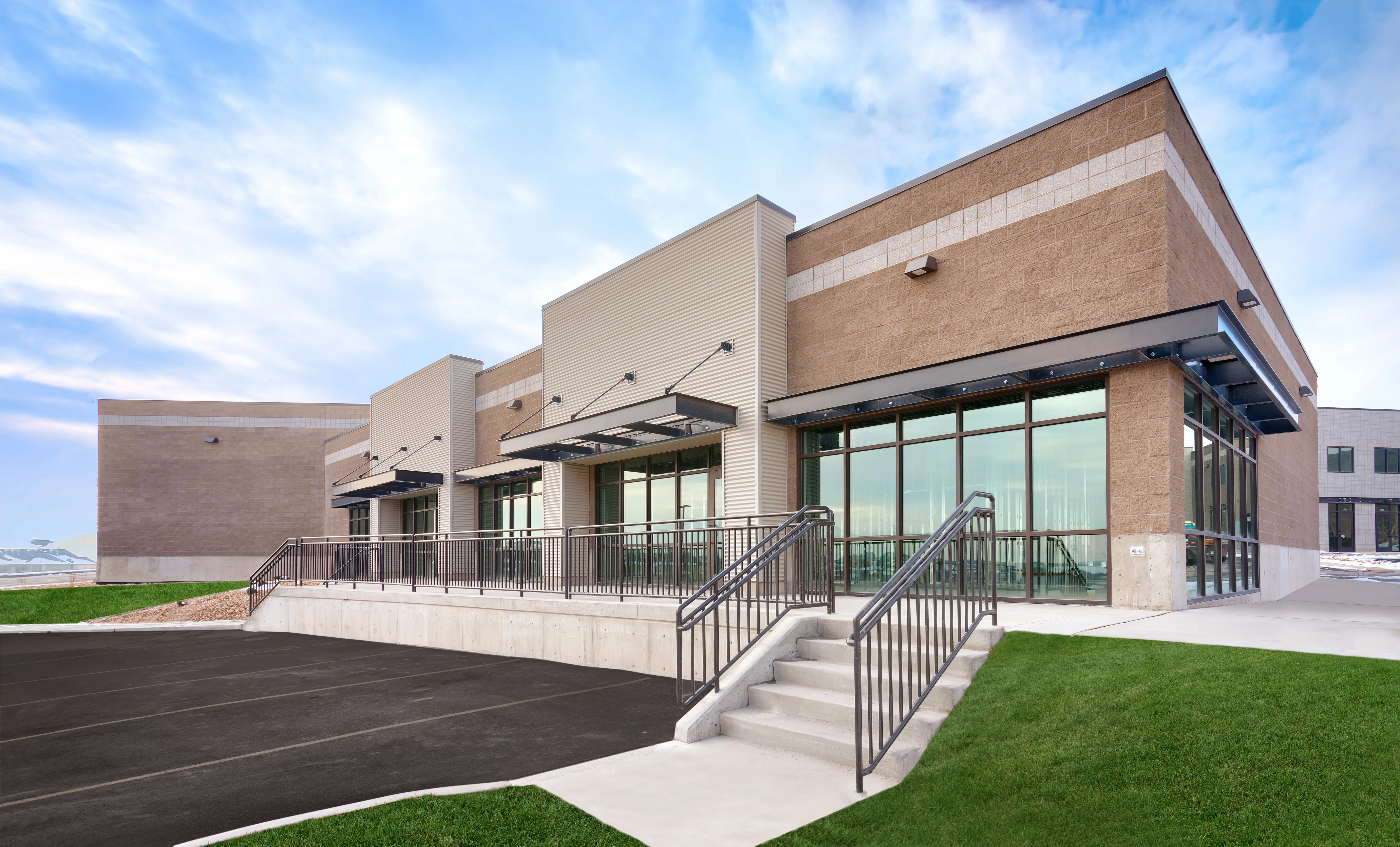 A wide-angle shot of a contemporary boutique office plaza with large floor-to-ceiling windows and dark metal industrial accents. A concrete staircase with minimalist metal handrails leads from a paved parking area to the elevated building entrance.