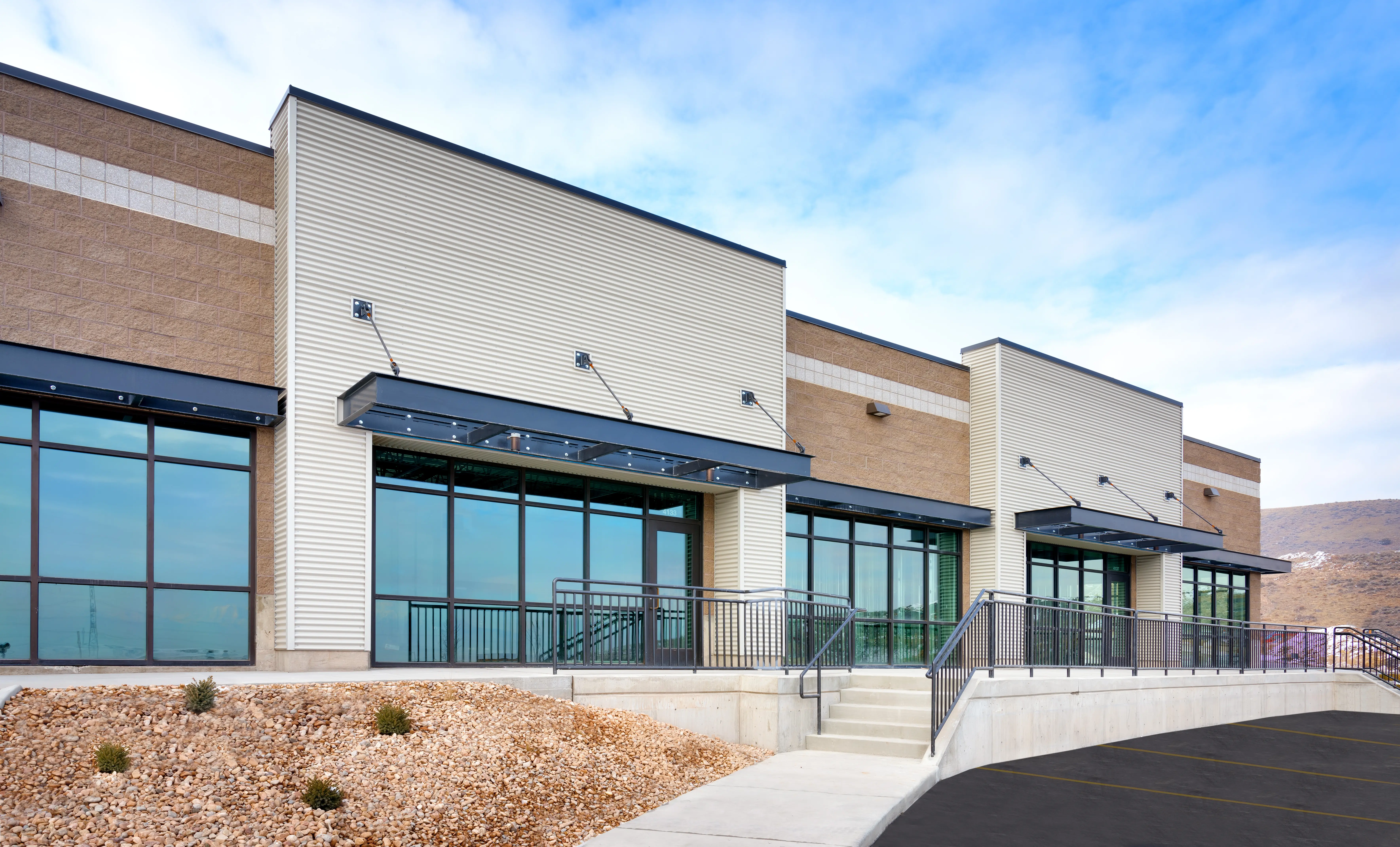 Close-up architectural view of a modern multi-unit commercial property under a soft blue sky. The facade showcases a mix of textured stone blocks, corrugated metal siding, and black-framed windows with cantilevered metal awnings over the entryways.