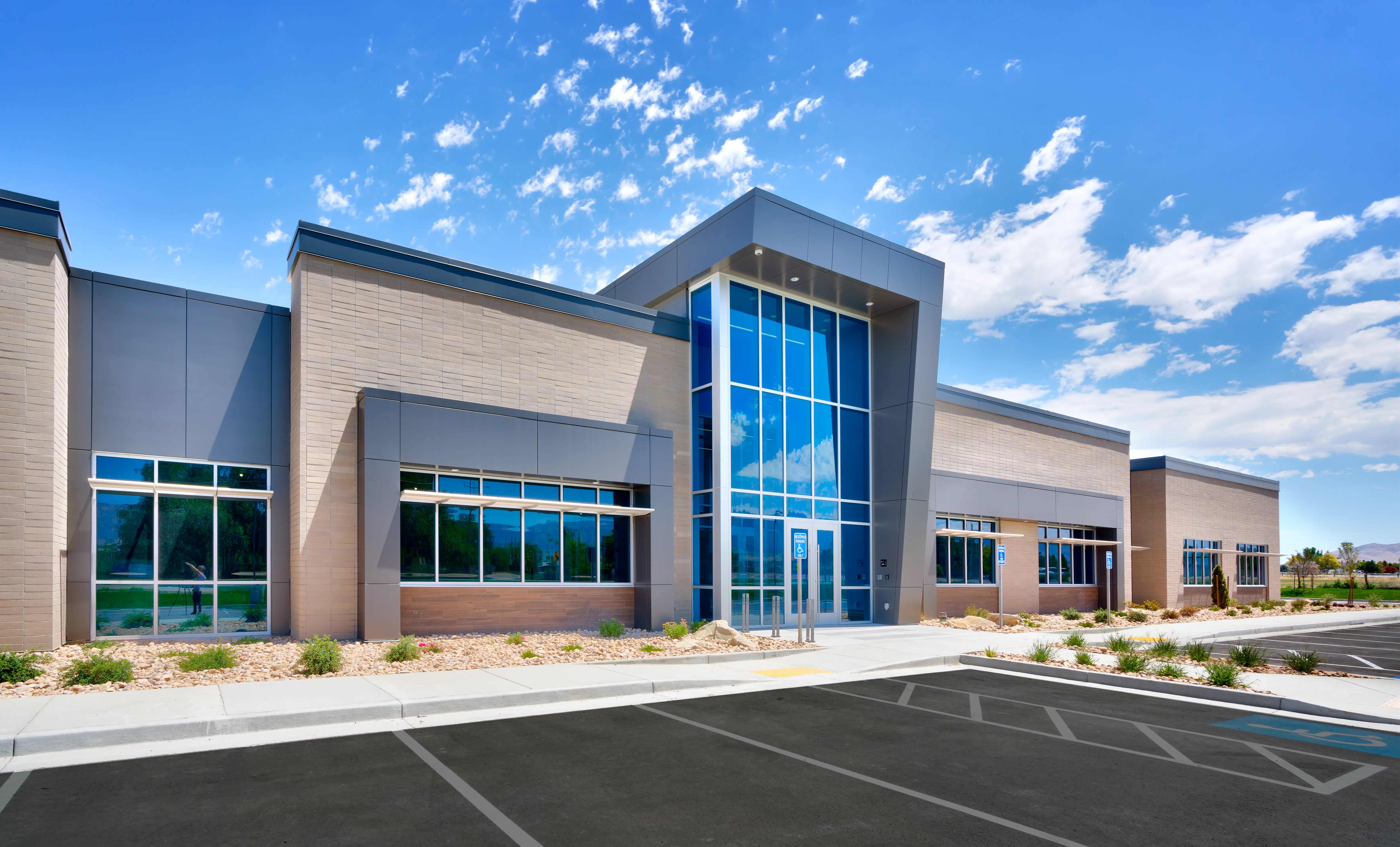 Modern commercial building with large glass windows, beige brick walls, and a parking lot under a blue sky.