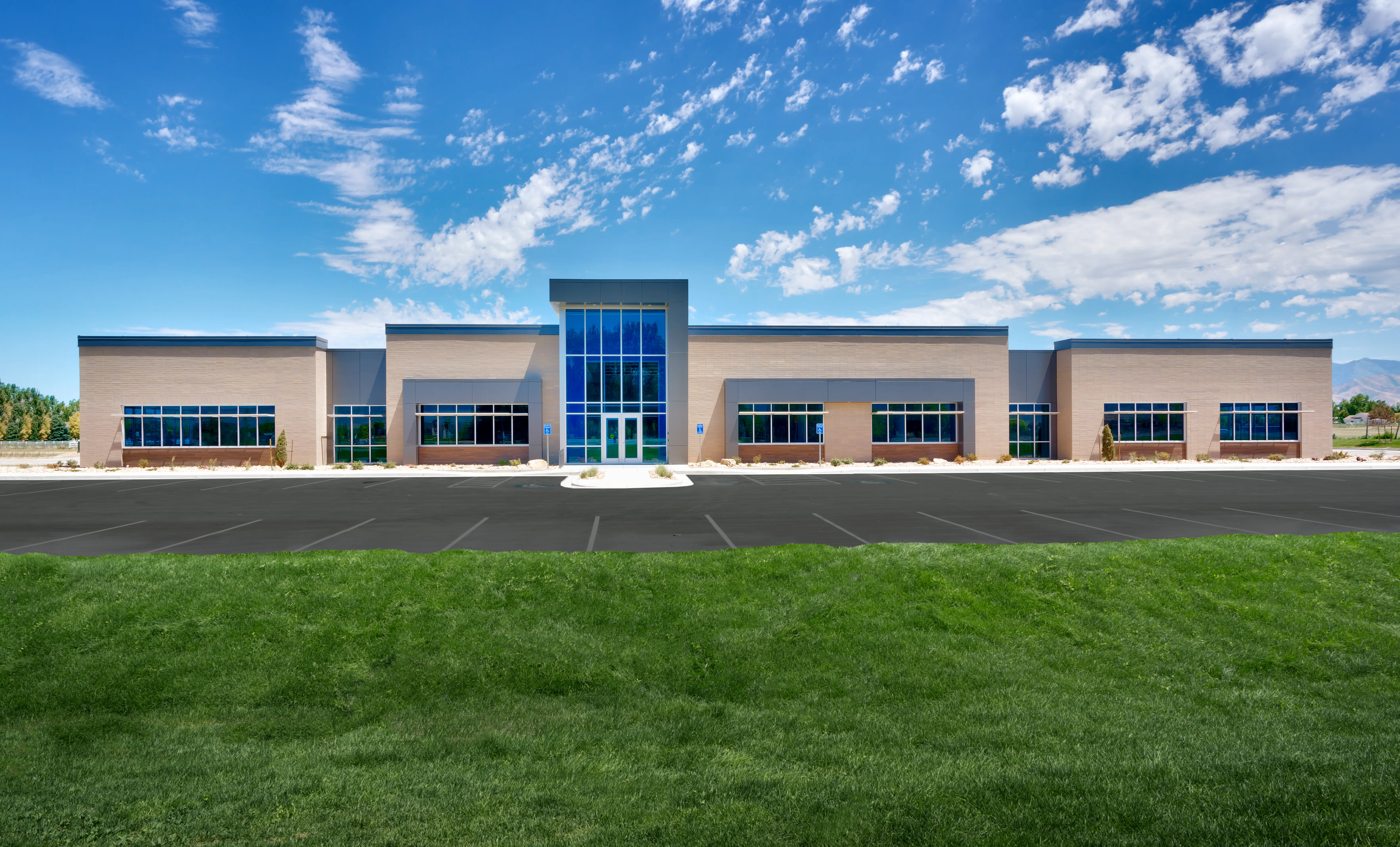 Modern one-story office building with large glass entrance, beige walls, and empty parking lot under a blue sky with scattered clouds.