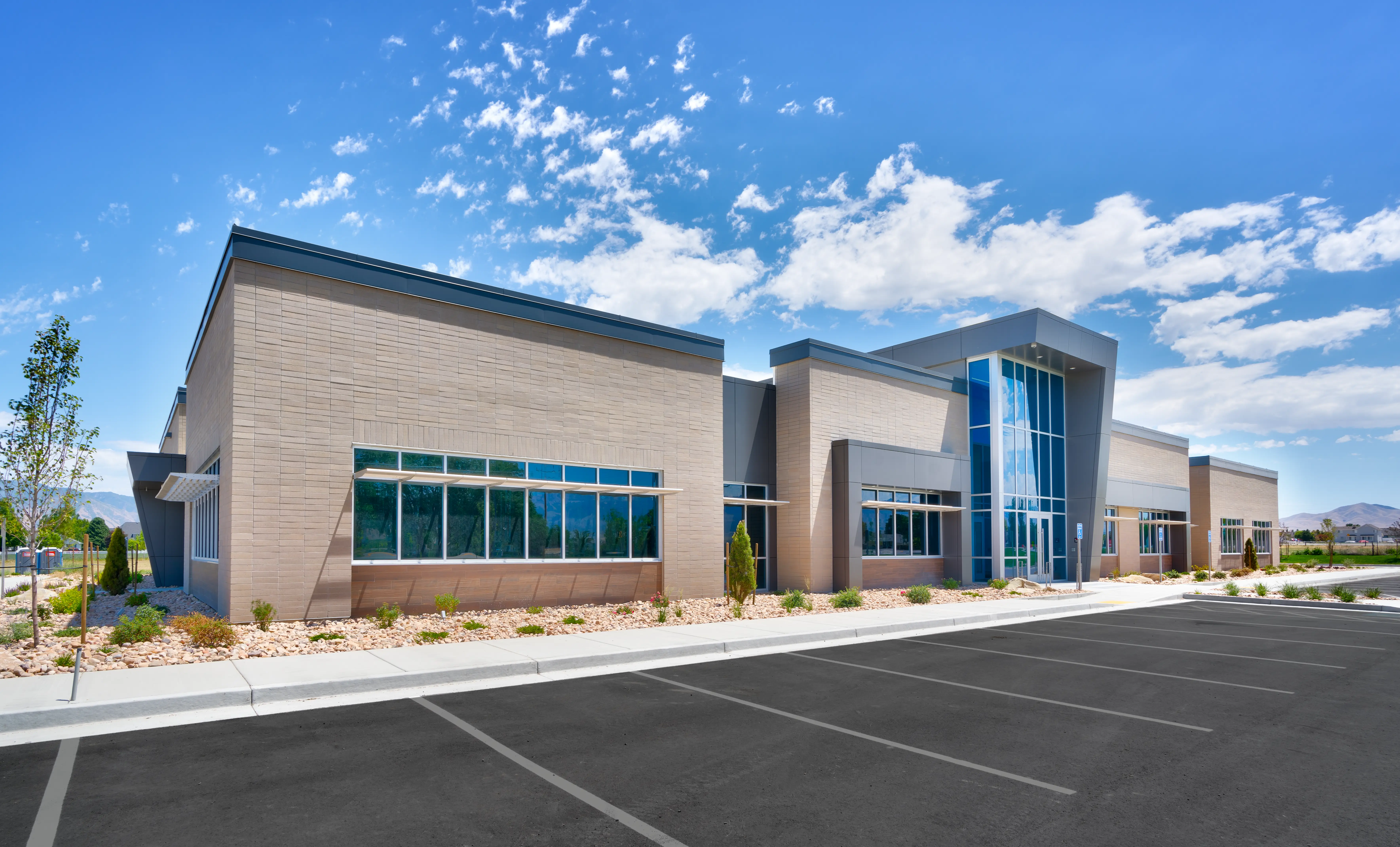 Modern single-story commercial building with large glass windows and a spacious empty parking lot under a blue sky with scattered clouds.