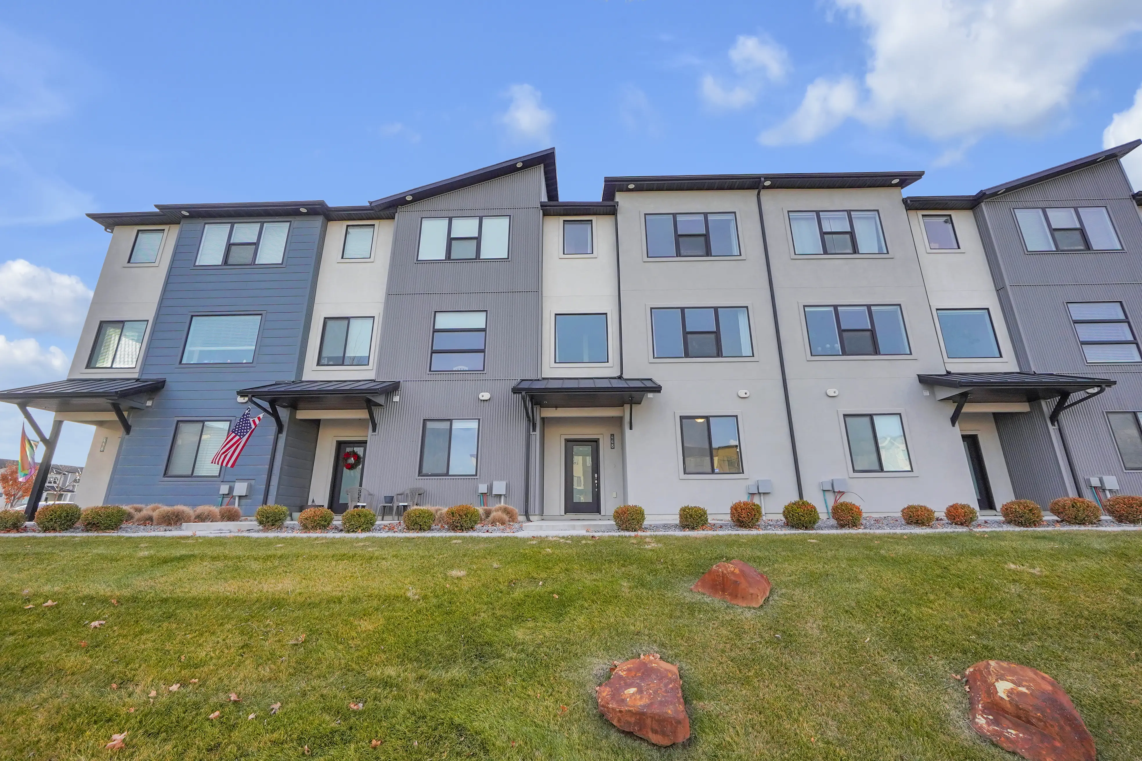 Modern three-story residential building with a green lawn and three large reddish rocks in front.