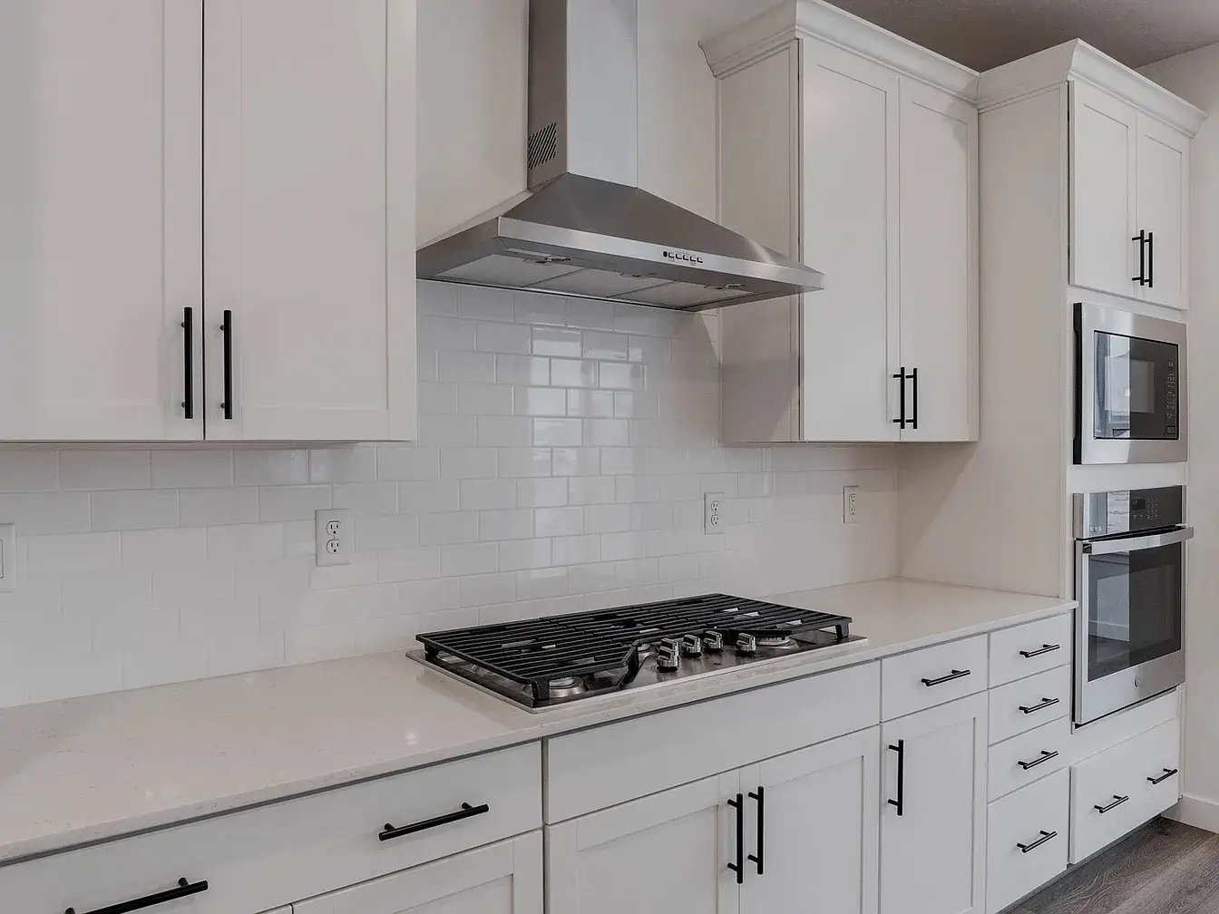Modern white kitchen with stainless steel gas stove, hood vent, microwave, and oven built into cabinets with black handles.