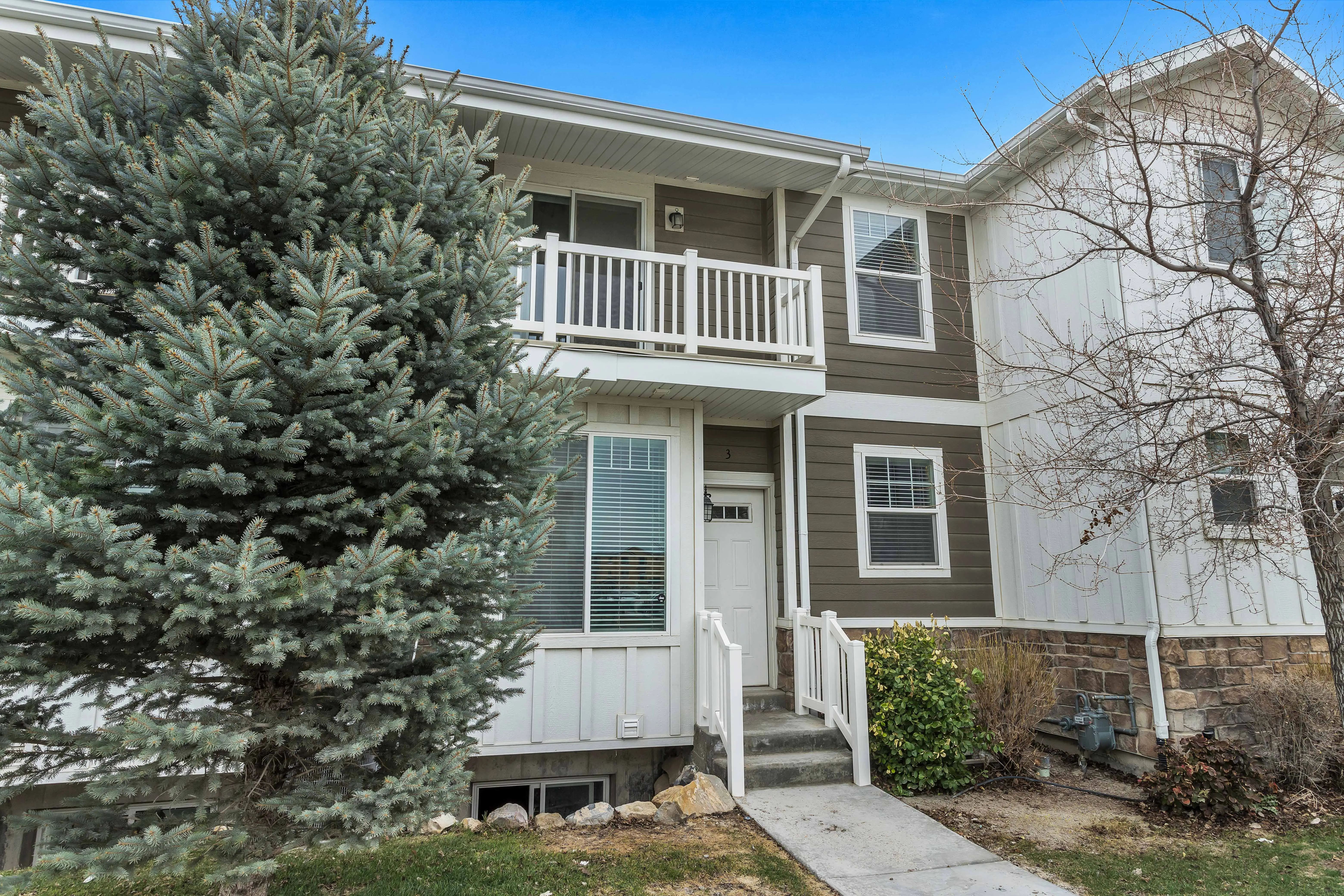 Two-story townhouse with white railings, brown and white siding, a concrete walkway, and a large evergreen tree in front.