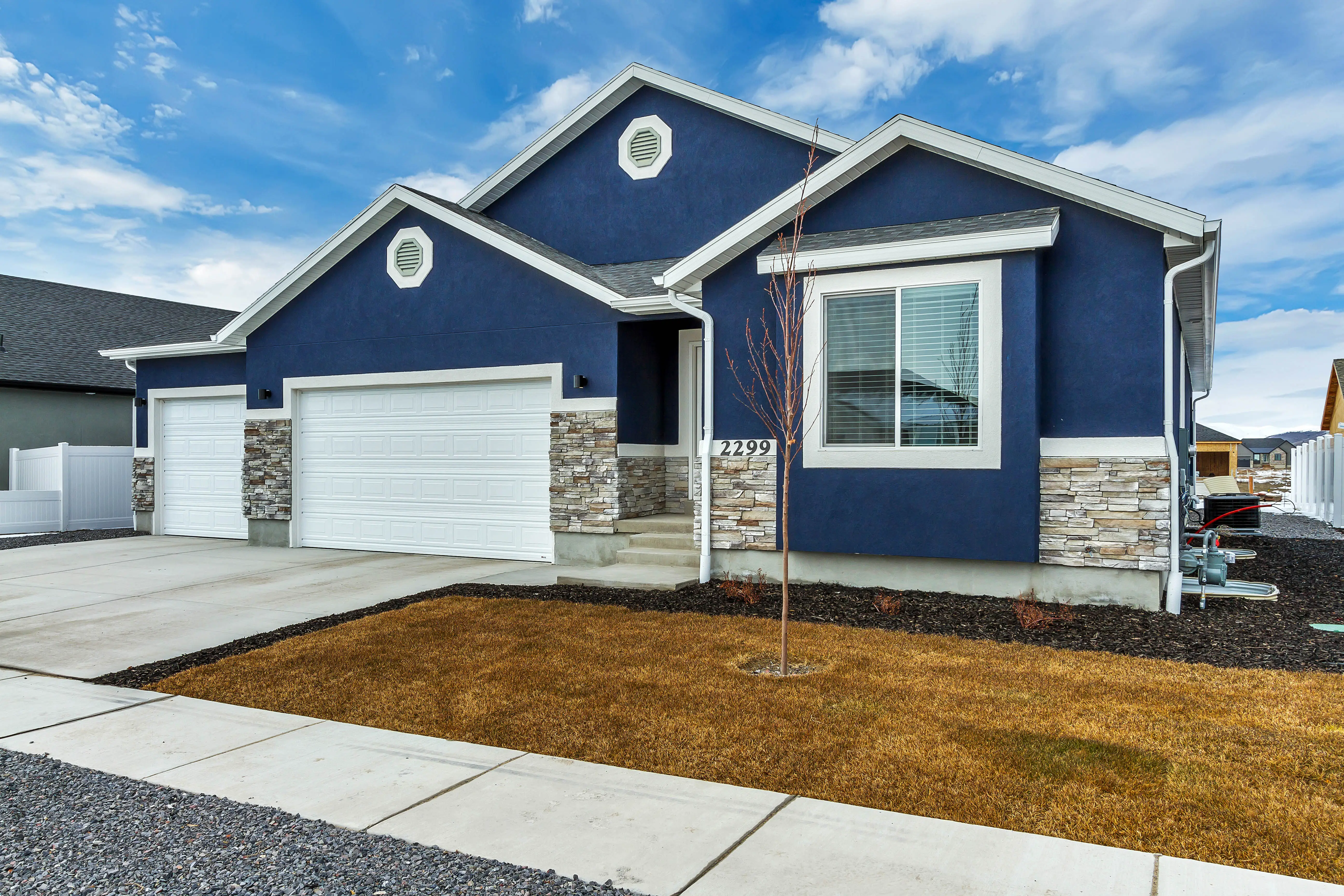 Modern single-story blue house with white trim, stone accents, double garage, and a small tree on a dry lawn.