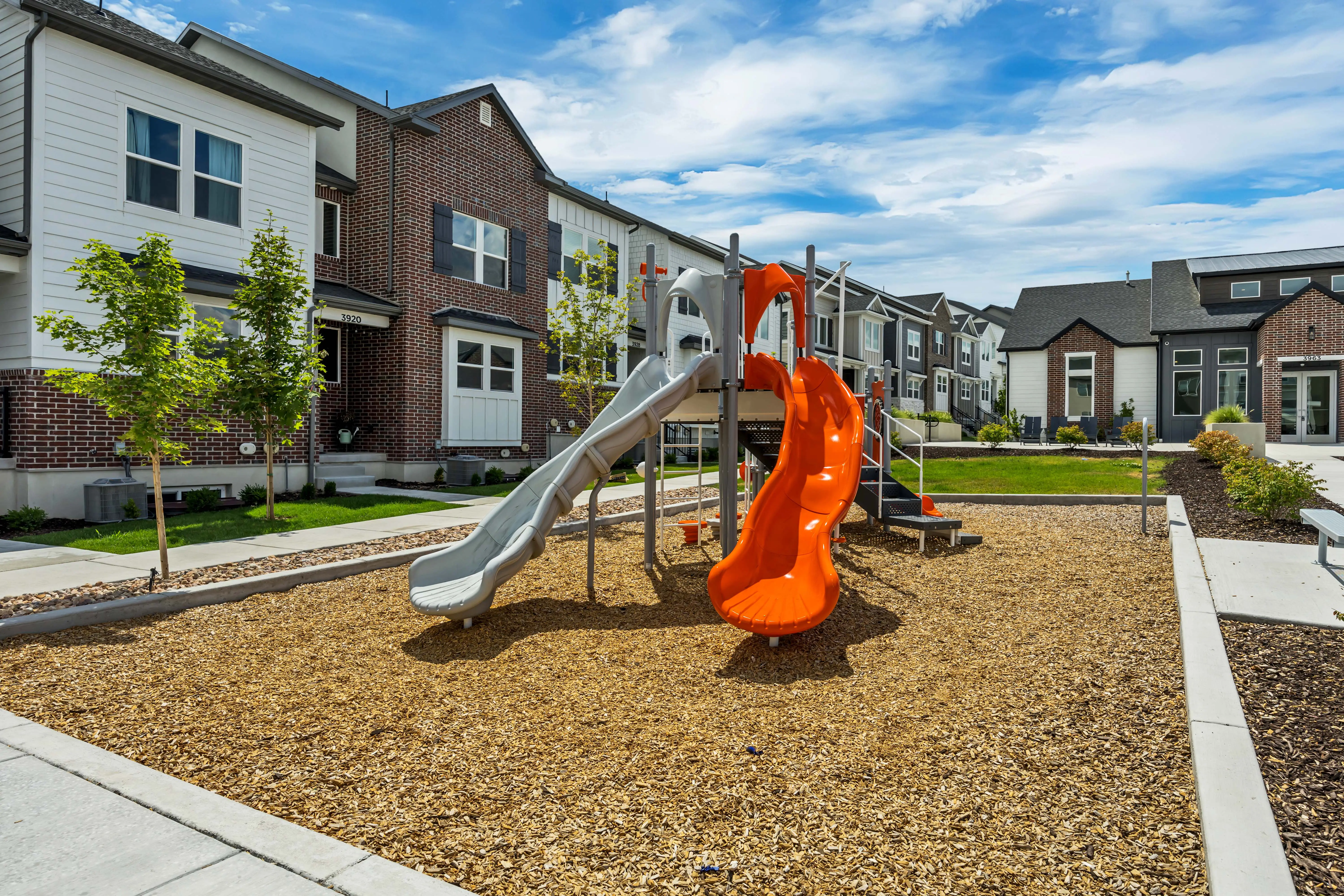 Playground with orange and gray slides on wood chip surface in front of modern townhouse buildings under a blue sky.