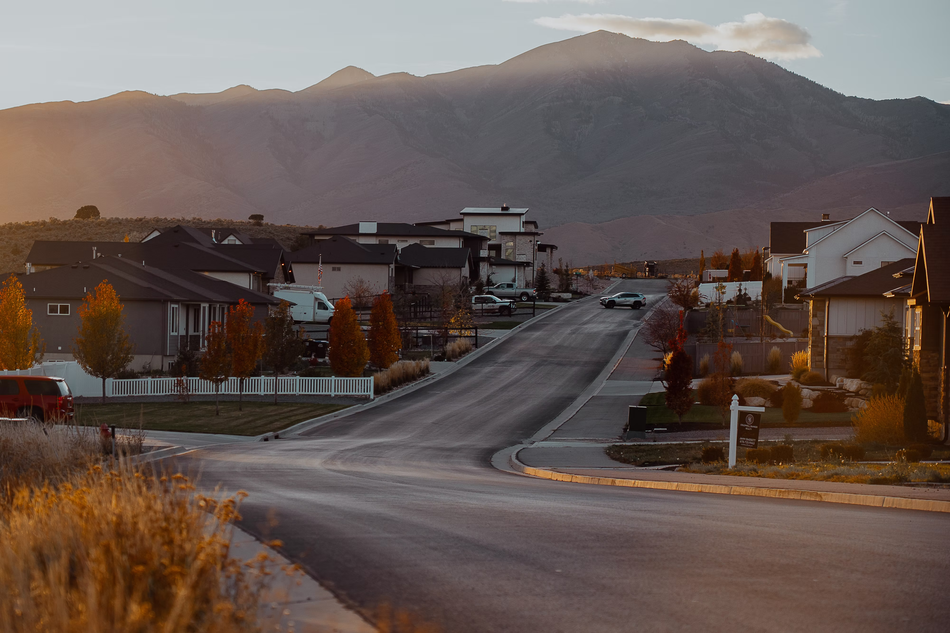 A quiet residential road curving through a high-end neighborhood at twilight, with the silhouetted peaks of a mountain range dominating the background.