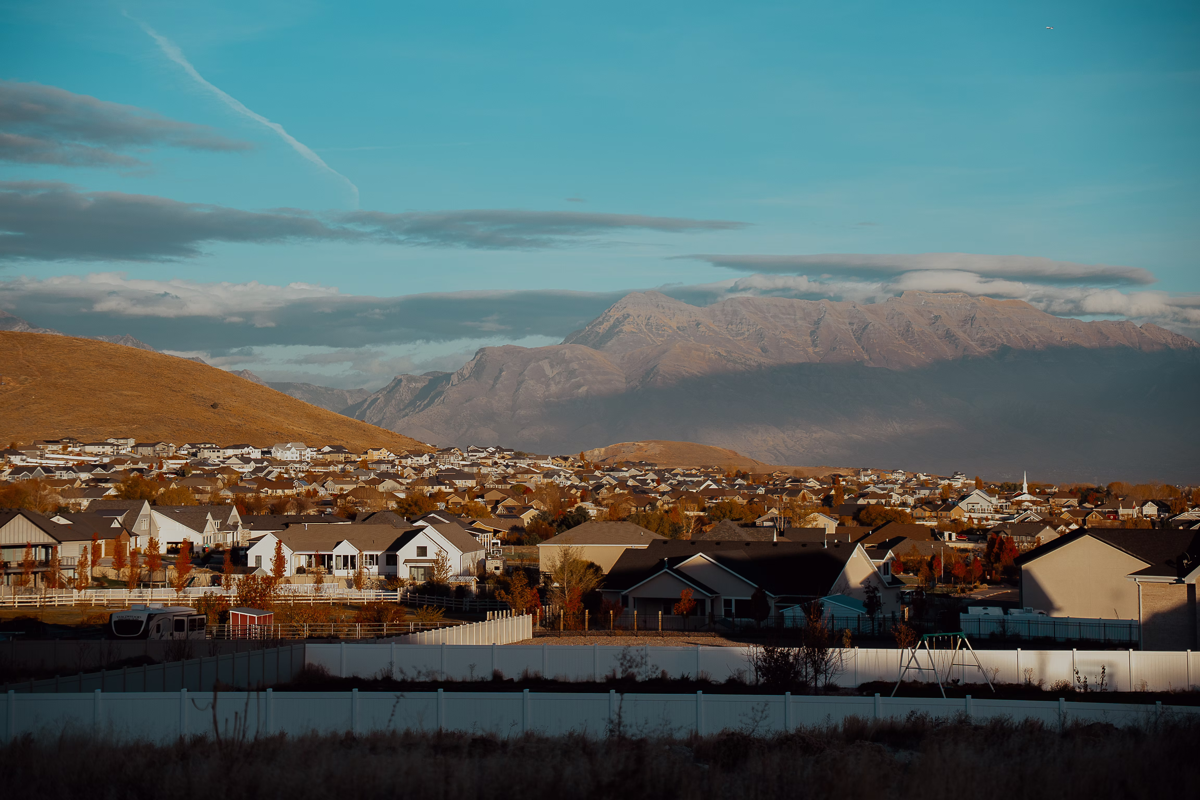 A wide-angle landscape view of a suburban valley neighborhood nestled at the base of expansive, rugged mountains under a clear blue sky.