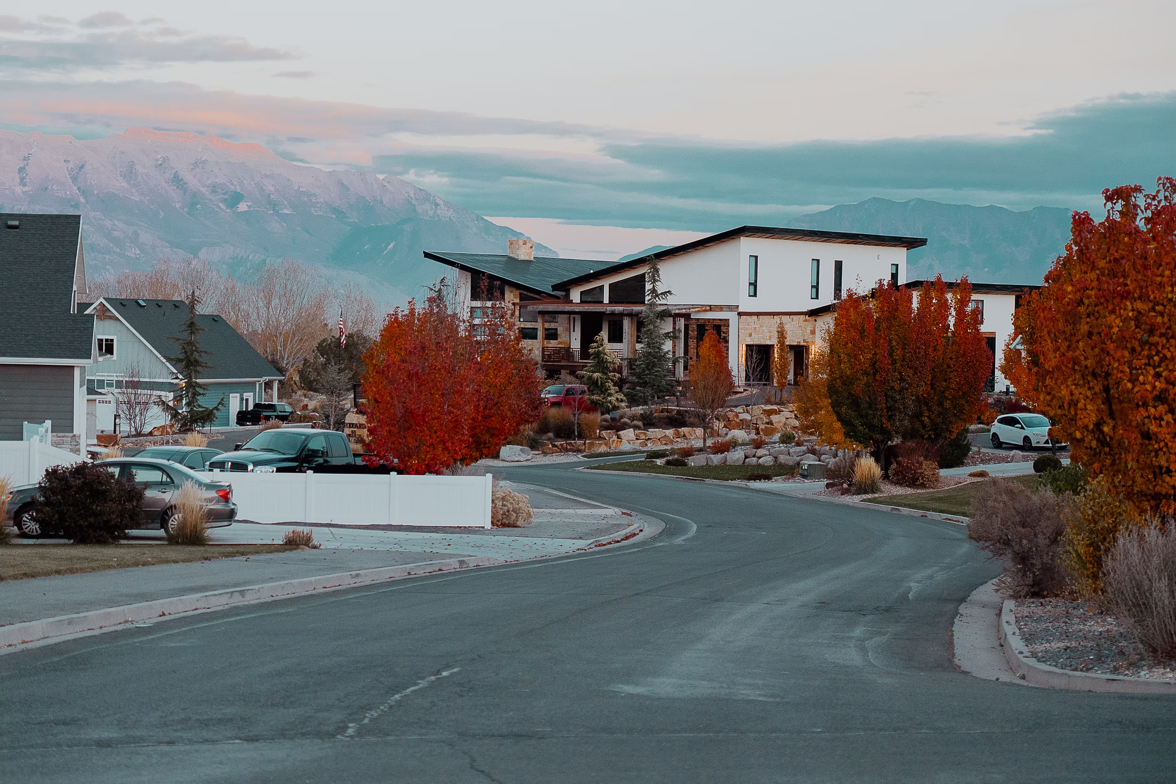A curved residential street leading toward a striking modern home with a unique slanted roofline, framed by vibrant red autumn trees and distant mountains.
