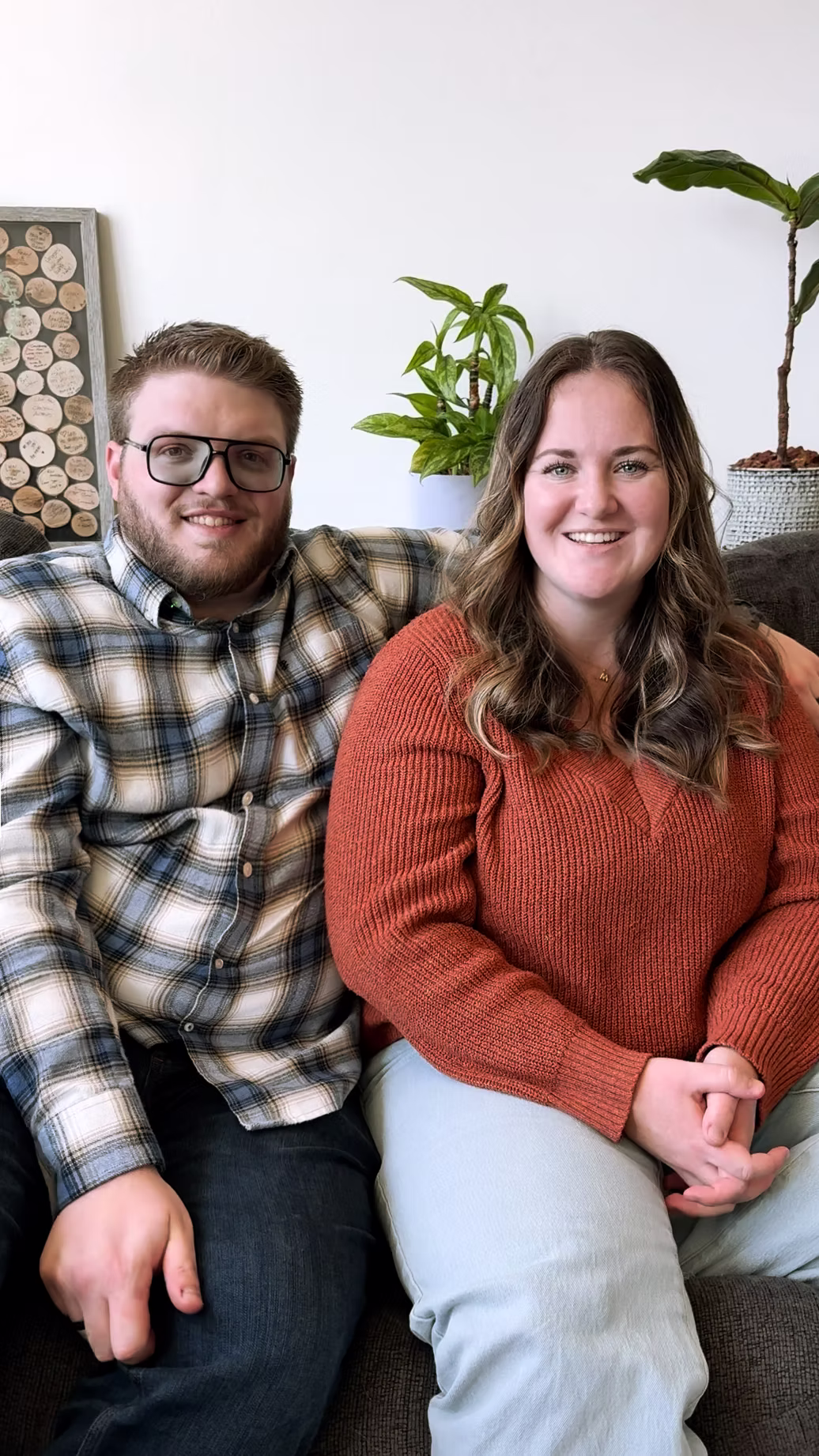Smiling man wearing glasses and plaid shirt sitting next to smiling woman in orange sweater on a couch with green plants in the background.
