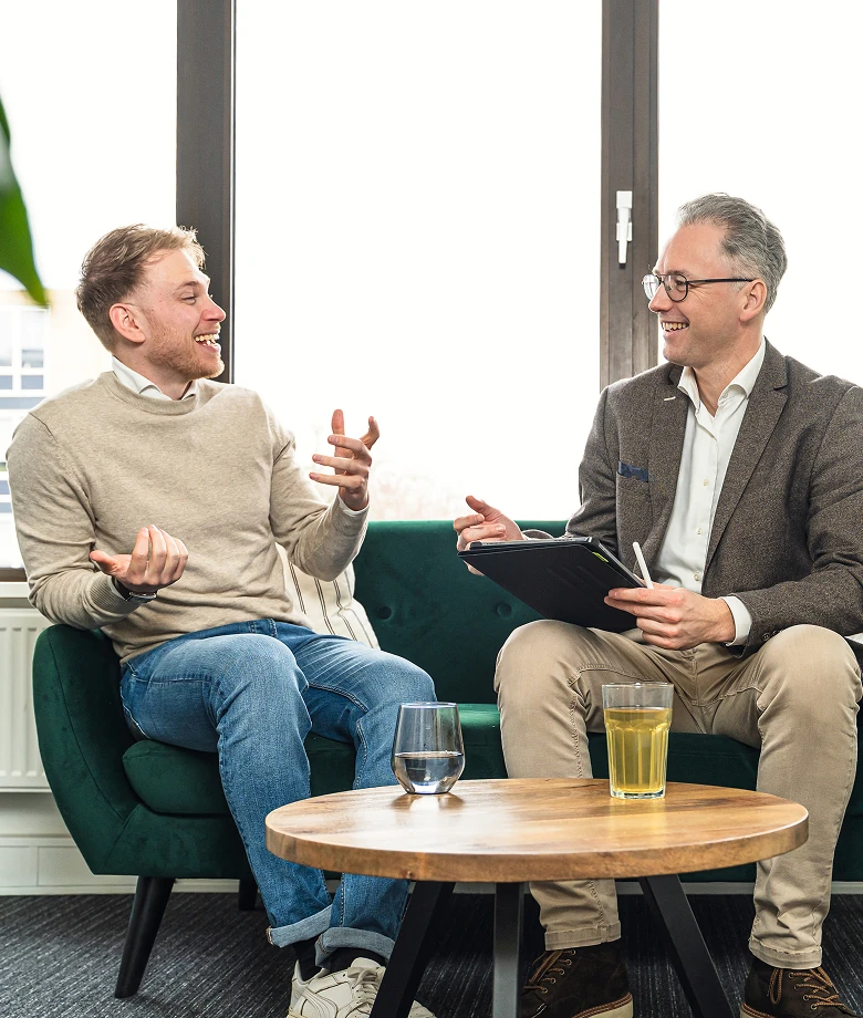Twee mannen zittend op een groene bank, pratend en lachend in een lichte kamer met een houten tafel met drankglazen ervoor.