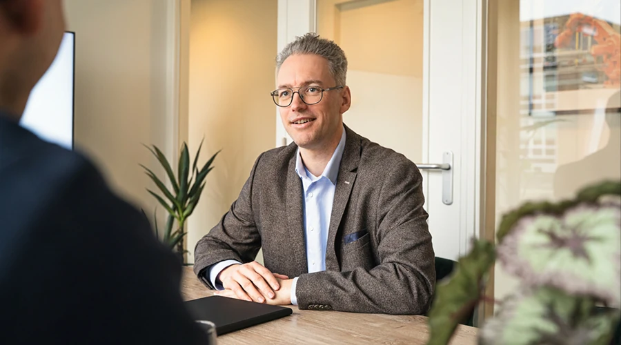 Man in bril en colbert zit aan tafel en voert een gesprek met een persoon tegenover hem, kantooromgeving met planten.