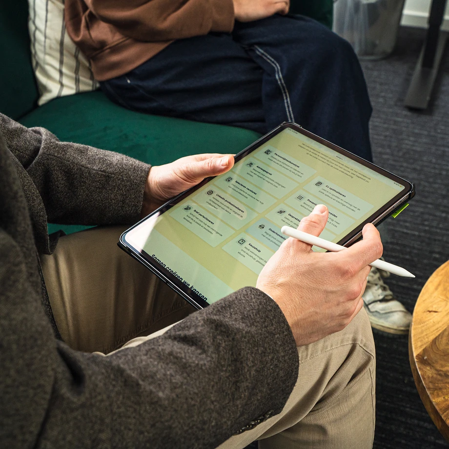 Persoon die een tablet vasthoudt en bedient met een stylus terwijl een ander op een bank zit.