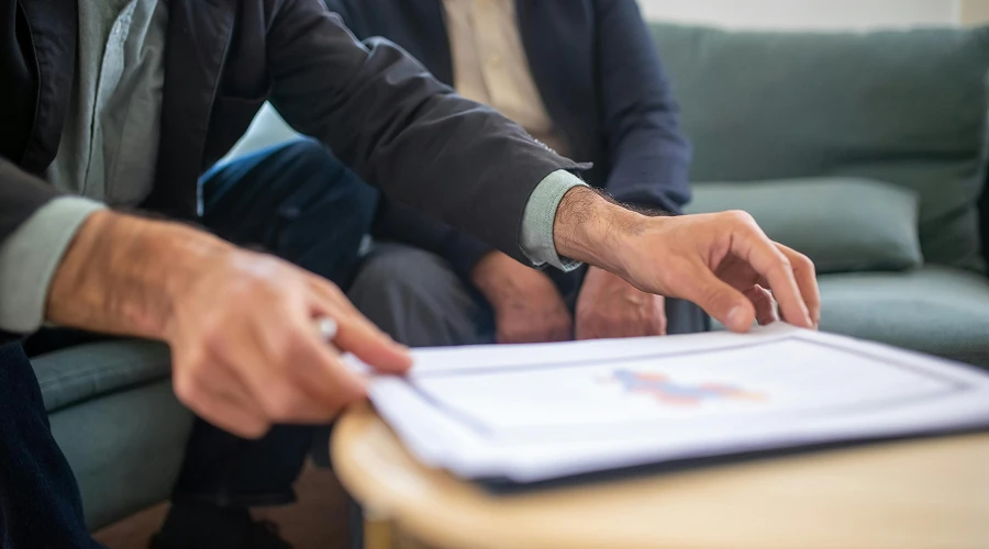 Twee personen zittend op een bank, waarbij een persoon met de hand door documenten bladert op een tafel.