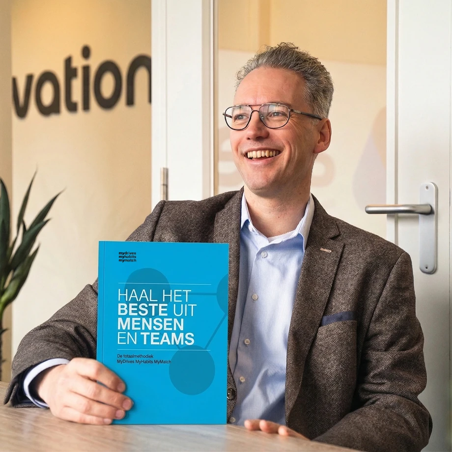 Man met bril en een bruin colbert glimlacht terwijl hij een blauw boek vasthoudt met de titel 'Haal het beste uit mensen en teams'.