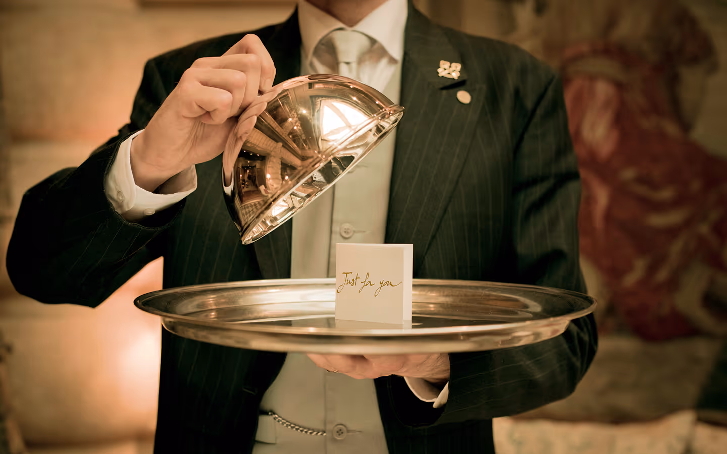 A waiter in formal attire lifts a silver cloche from a tray revealing a card that reads 'Just for you'.