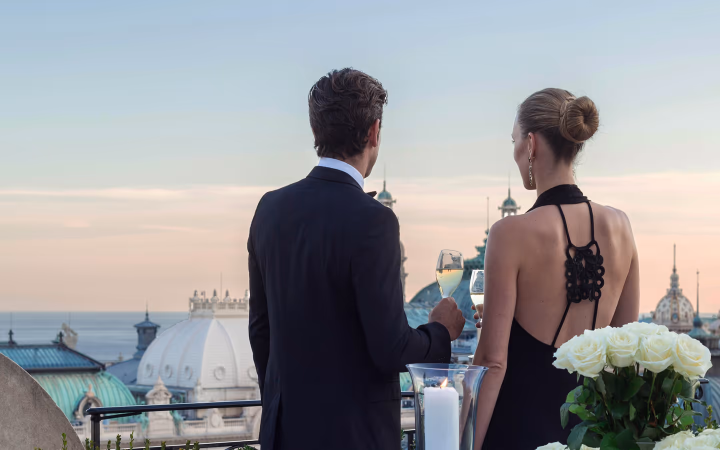 Well-dressed man and woman holding wine glasses, standing on a terrace overlooking a city skyline with domed rooftops at sunset.