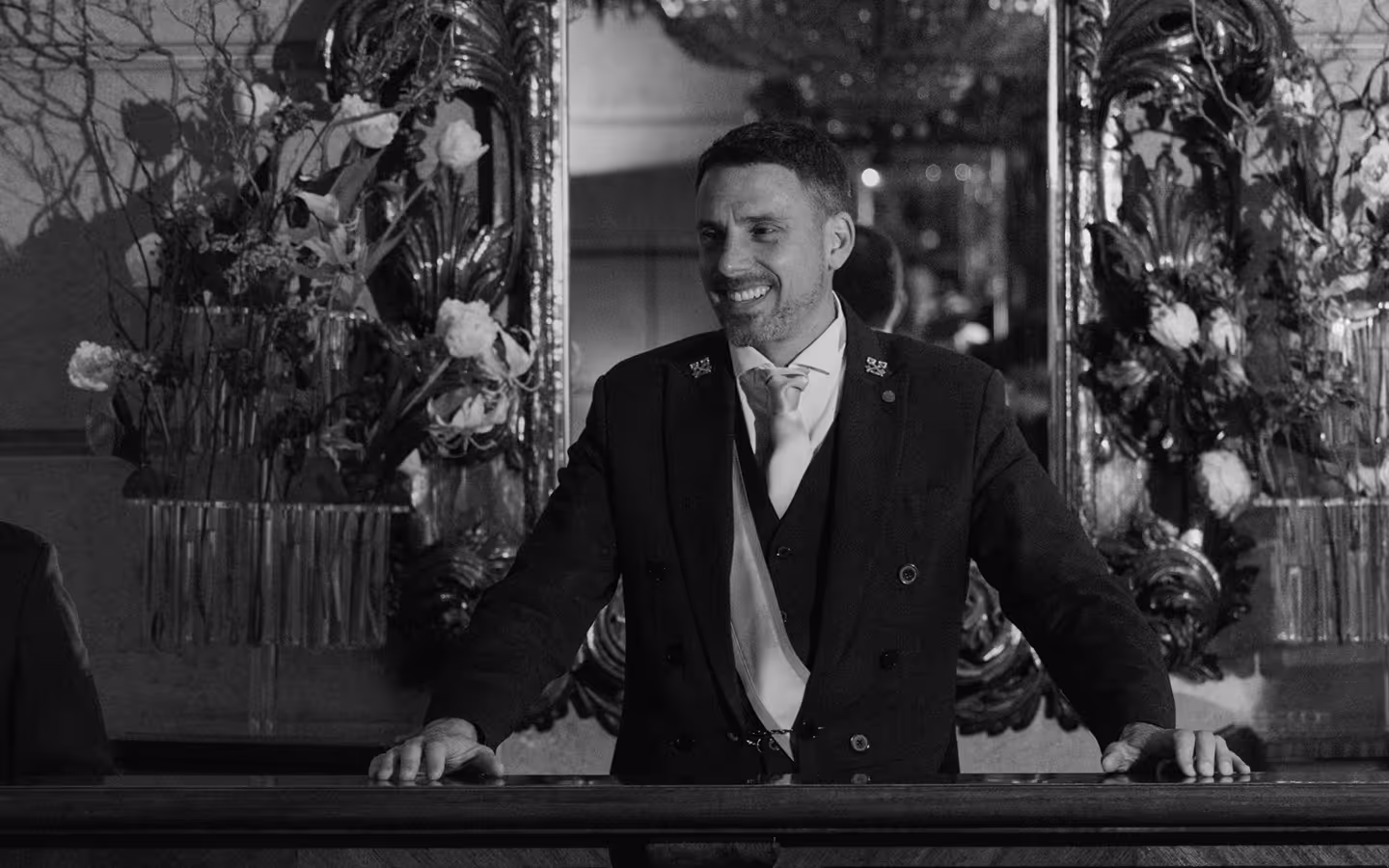Smiling man in formal attire leaning on a polished wooden counter with floral arrangements and ornate mirrors in the background.
