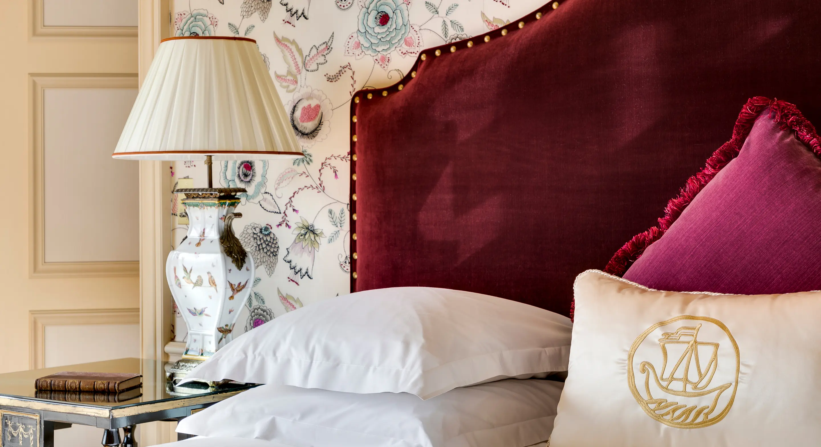 Elegant bedroom corner with a red velvet headboard, white pillows, decorative cushion, floral wallpaper, and a vintage table lamp on a glass side table.