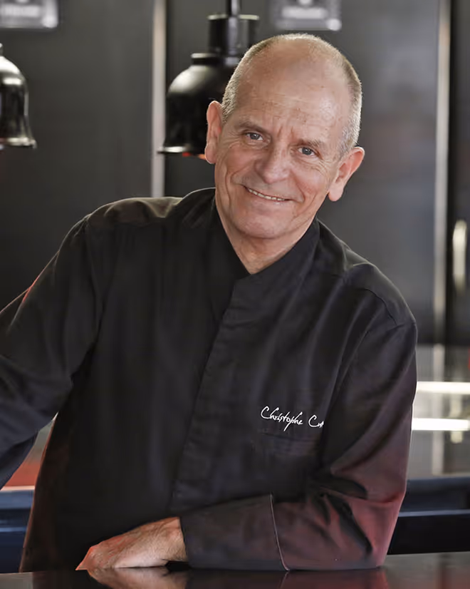 Smiling middle-aged man in a black chef coat leaning on a counter in a modern kitchen.