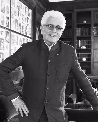 Elderly man with white hair and glasses standing indoors in a room with bookshelves and framed portraits on the wall.
