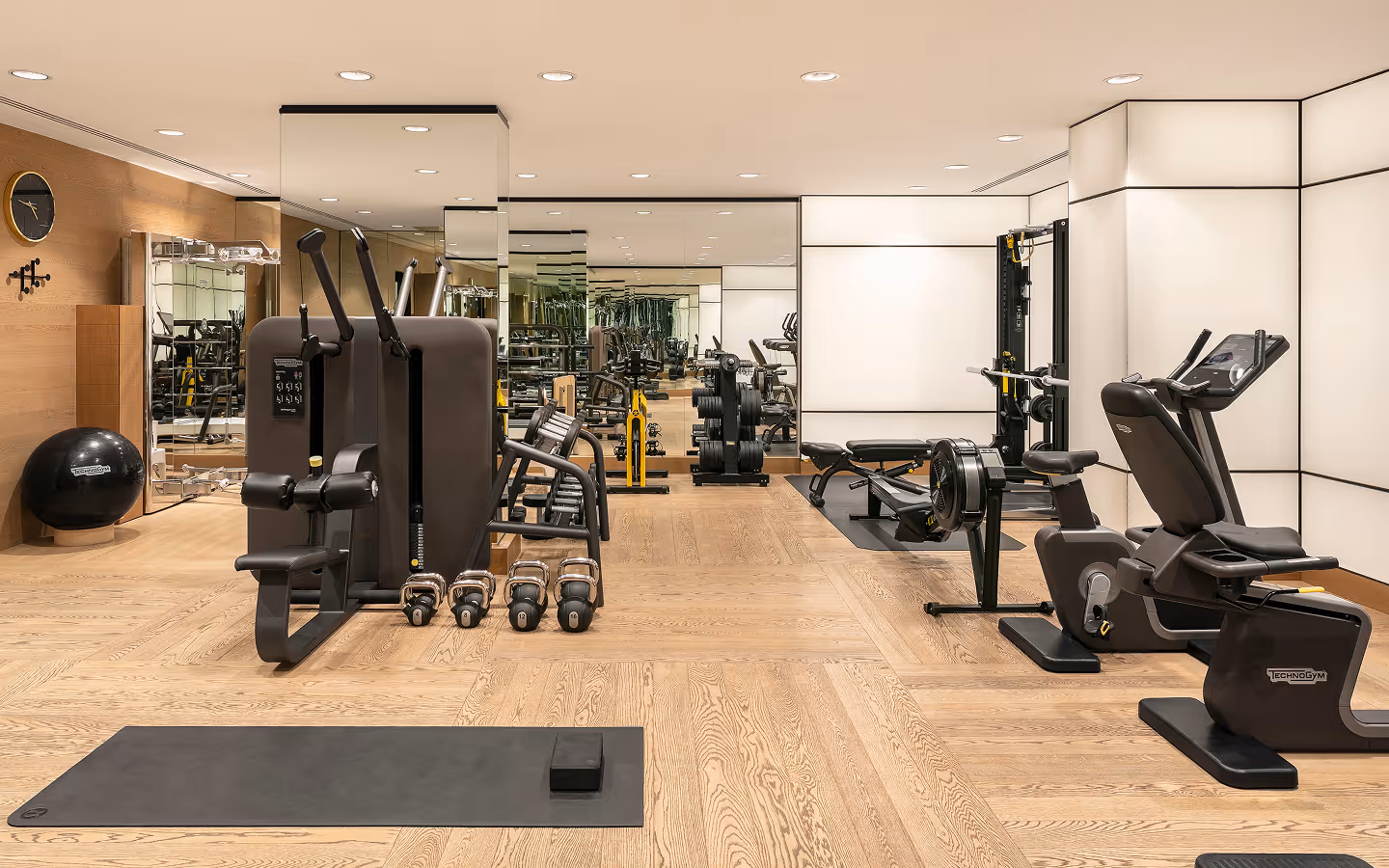 Modern gym room with wooden floors, various workout machines, kettlebells, and a yoga mat.
